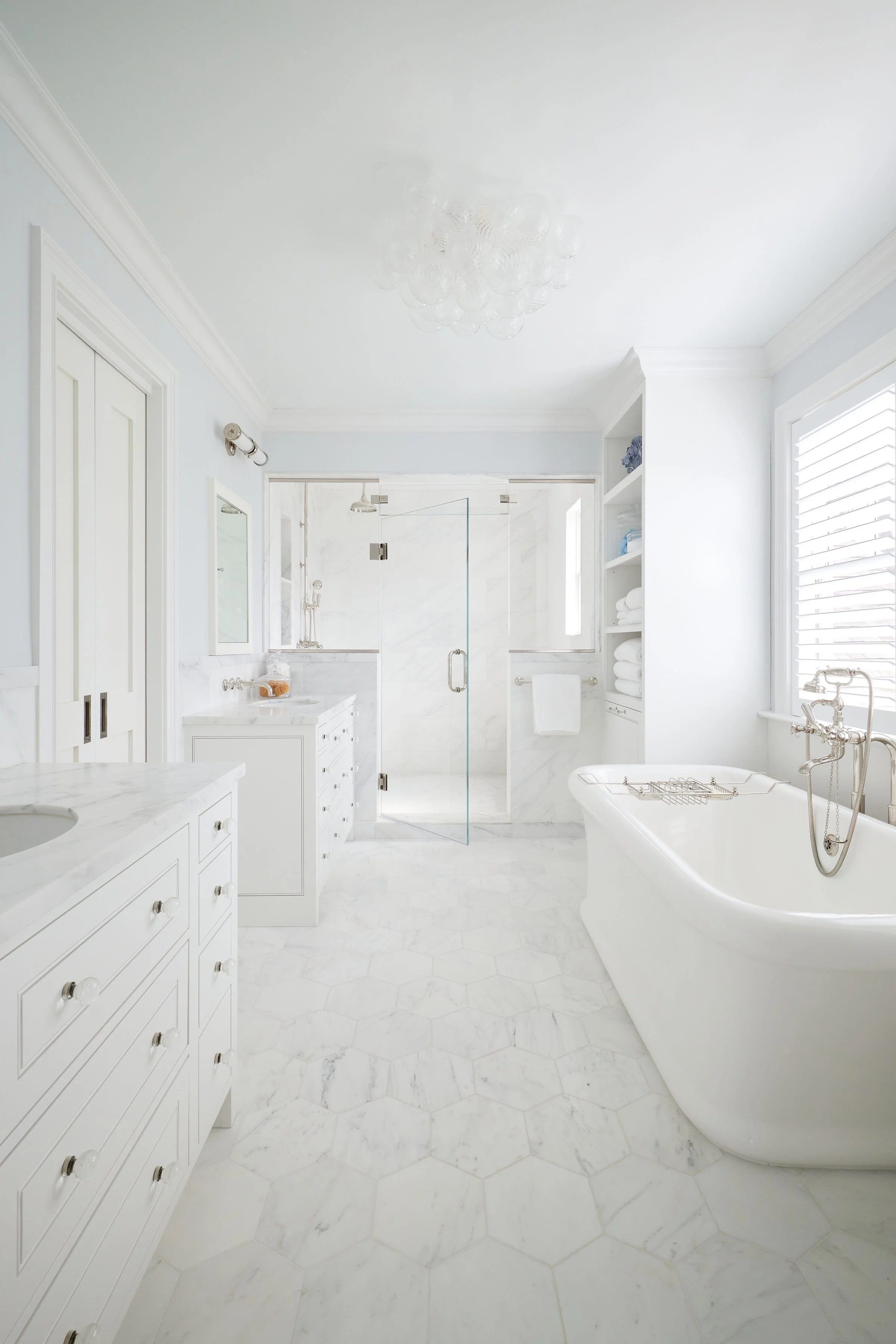 Bright, white bathroom with marble floors and walls, featuring a vintage bathtub with a silver faucet, a glass-enclosed shower, and built-in shelving filled with towels and toiletries.