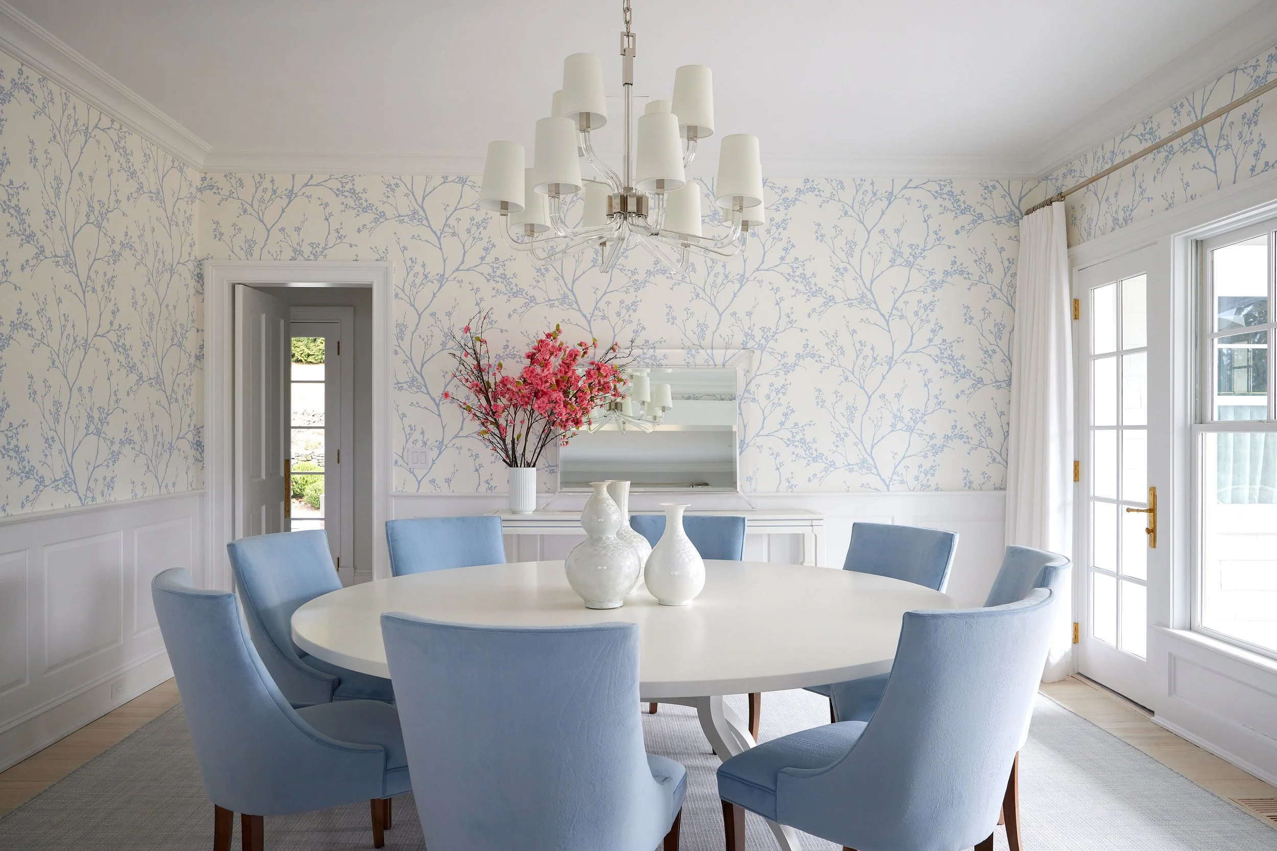 A dining room with a round white table surrounded by light blue upholstered chairs. The room has wallpaper with a blue and white tree branch pattern. There are white vases with pink flowers on the table. A chandelier with multiple white lampshades hangs from the ceiling, and large windows with white curtains let in natural light.