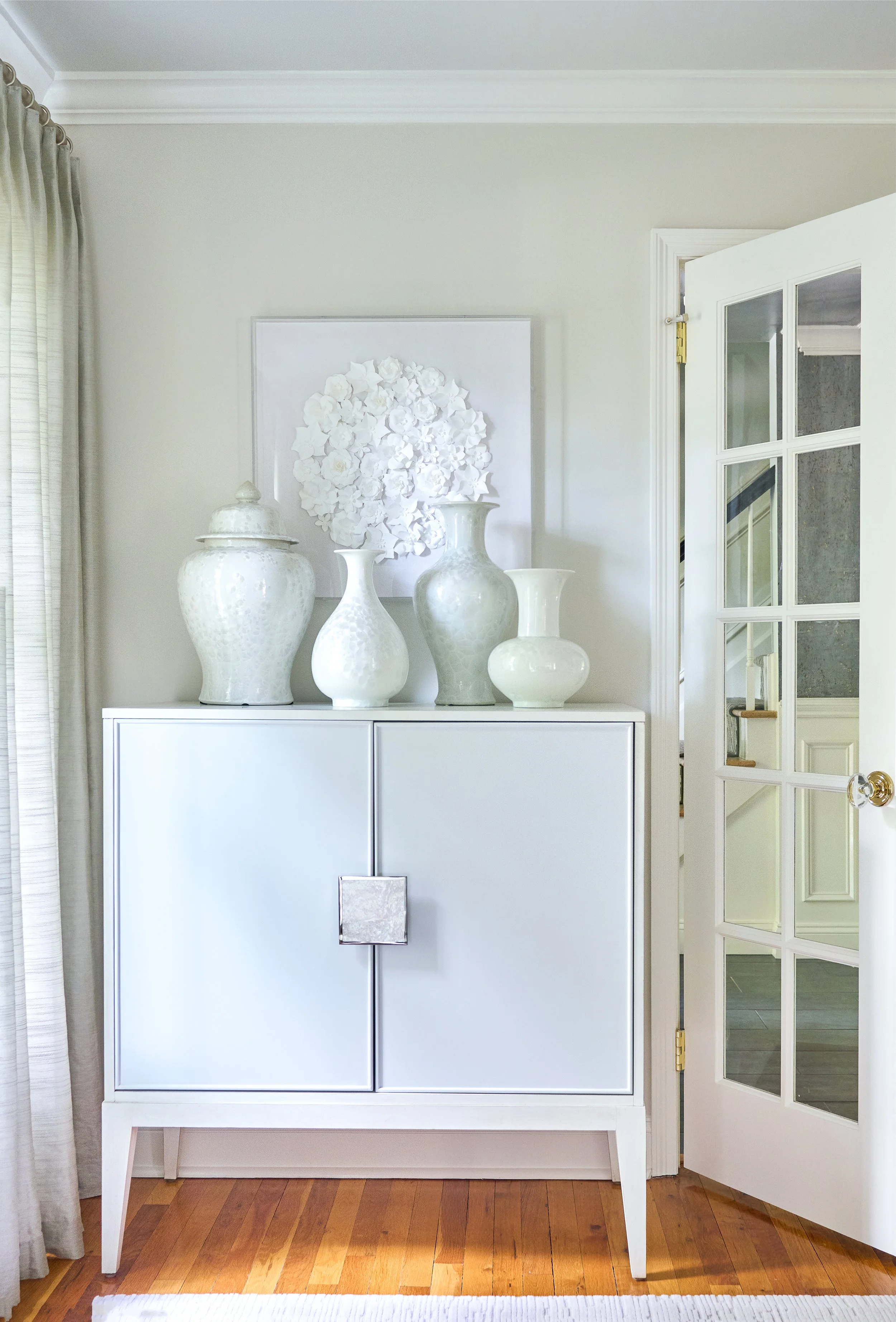 White cabinet with four vases with textured surfaces on top, artwork with white floral design on the wall, French door to the right, hardwood floor, and partial window with beige curtains on the left.