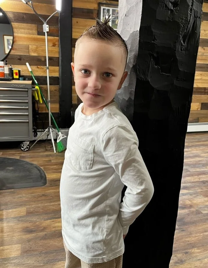 Young boy with spiky hair wearing a white long-sleeve shirt, standing in a room with wooden wall decor, a mirror, and cleaning supplies in the background.