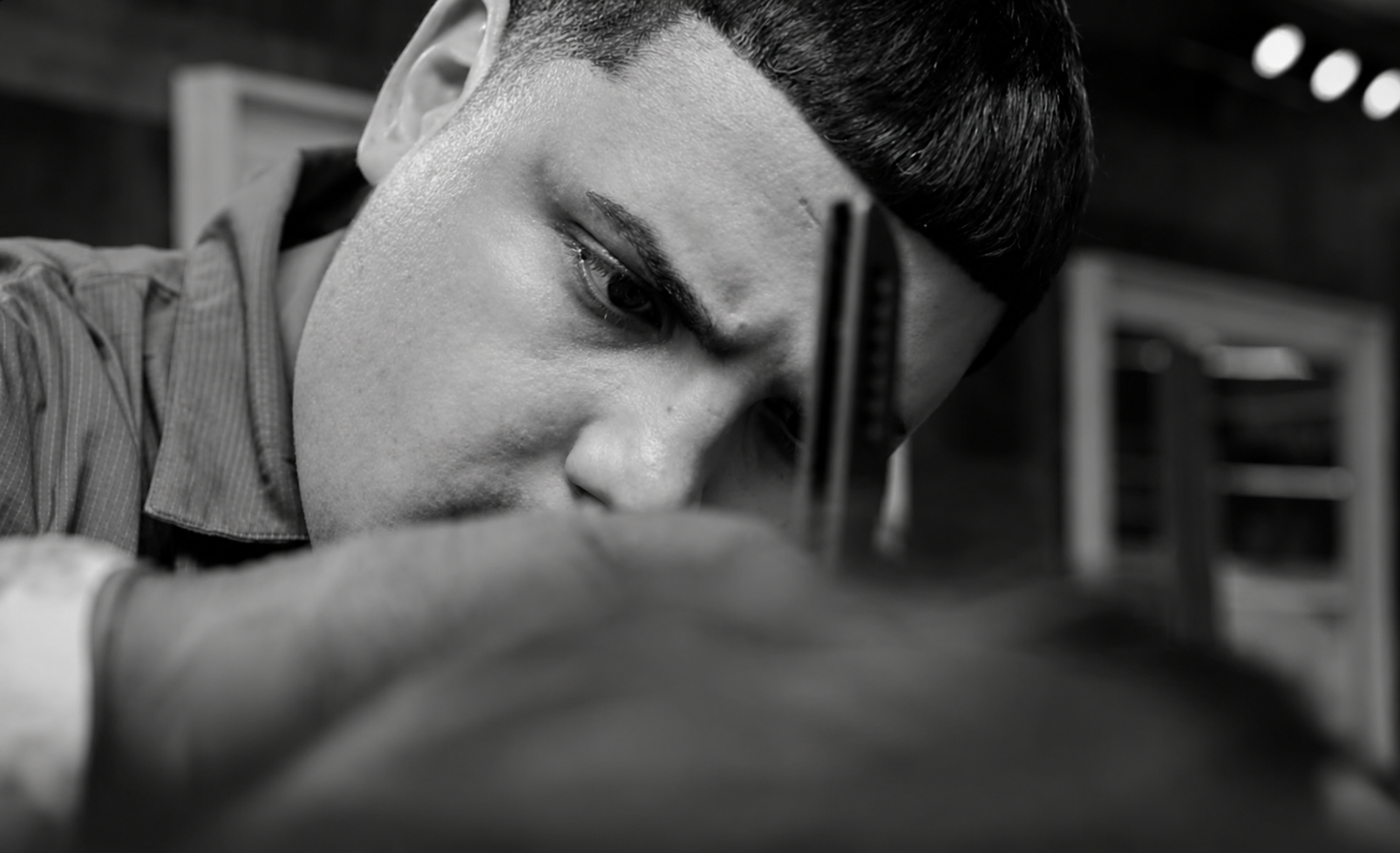 A young man with a haircut focused intently on woodworking or crafting with a sharp tool in his hand in a workshop.