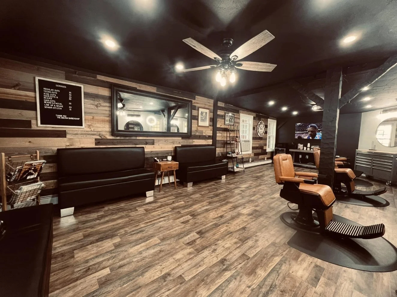 Interior of a barbershop with barber chairs, a wooden wall, a TV, and a service menu board.