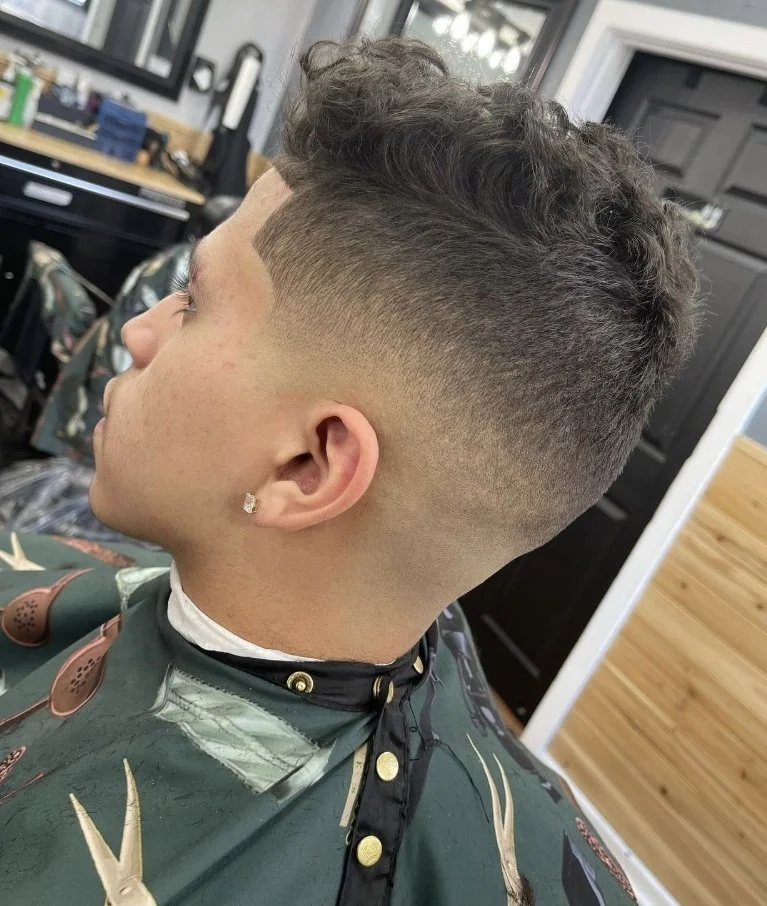 Young man with a modern fade haircut sitting in a barbershop chair, wearing a barber cape with barber tools background.