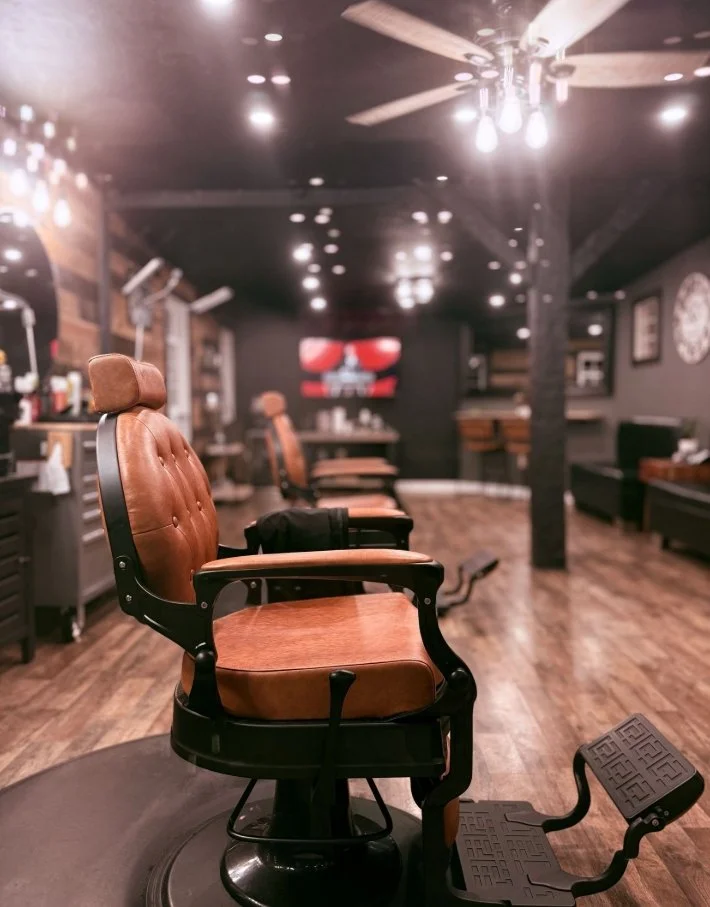 A vintage-style barbershop chair with brown leather upholstery and black frame in an empty barbershop with wood flooring and wall decor, illuminated by ceiling lights.