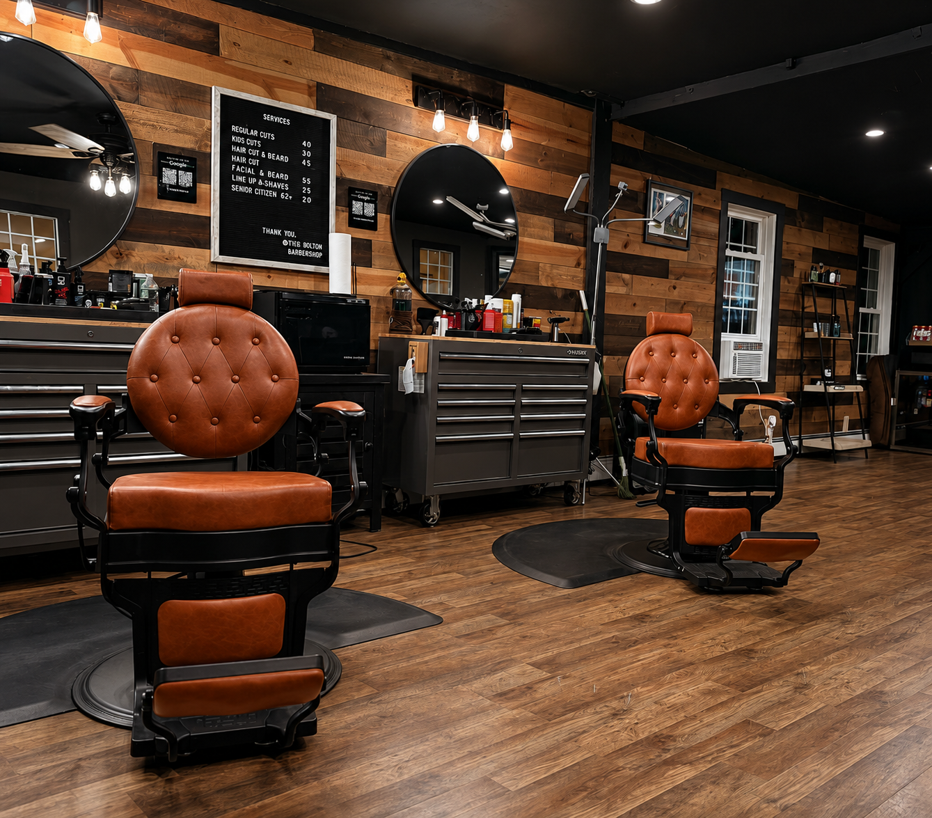 Interior of a barbershop with two vintage-style barber chairs with brown leather cushions, wooden armrests, and footrests, positioned on black round mats. The background features wooden wall panels, large round mirrors, and a black barber station with grooming tools and supplies. There are three windows with white frames, a shelving unit with products, and a black digital menu board listing services and prices.