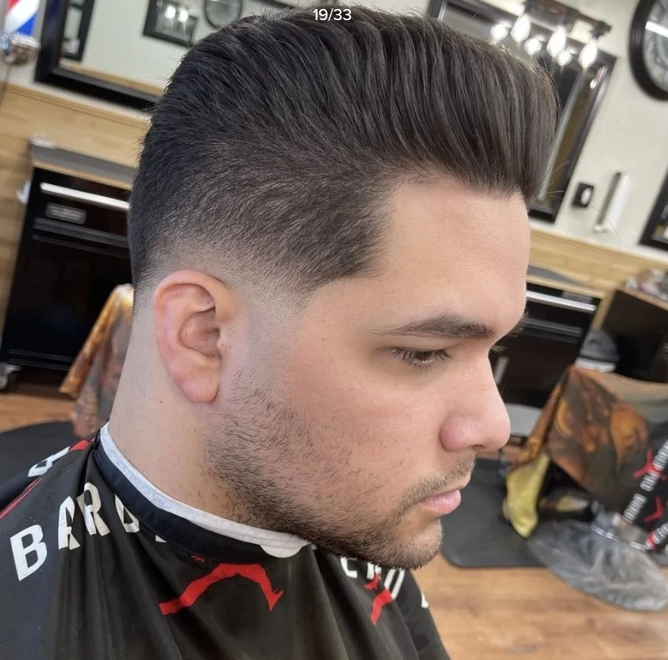 Close-up of a man with a fade haircut, styled slicked back, sitting in a barber shop.