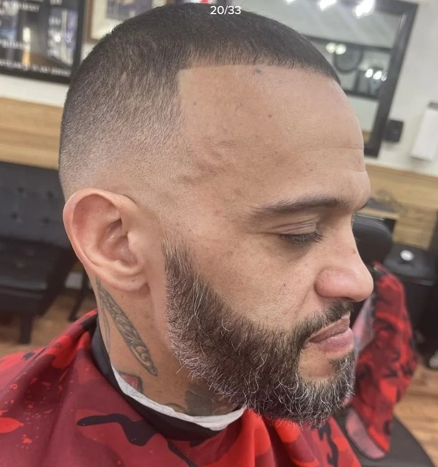 Side view of a man with a fresh fade haircut and a full salt-and-pepper beard in a barbershop.