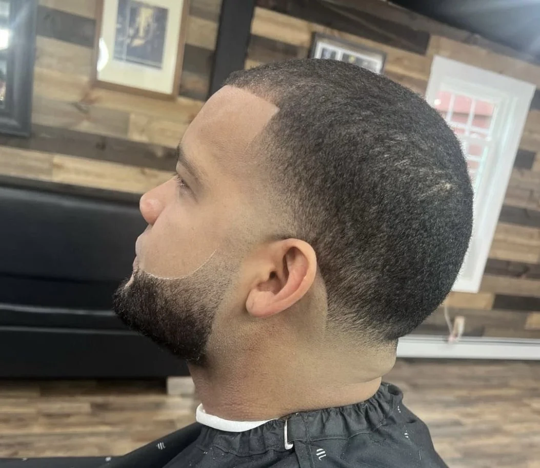 Side view of a man with a fresh haircut in a barbershop, featuring a fade with a line design, a beard, and a textured top.