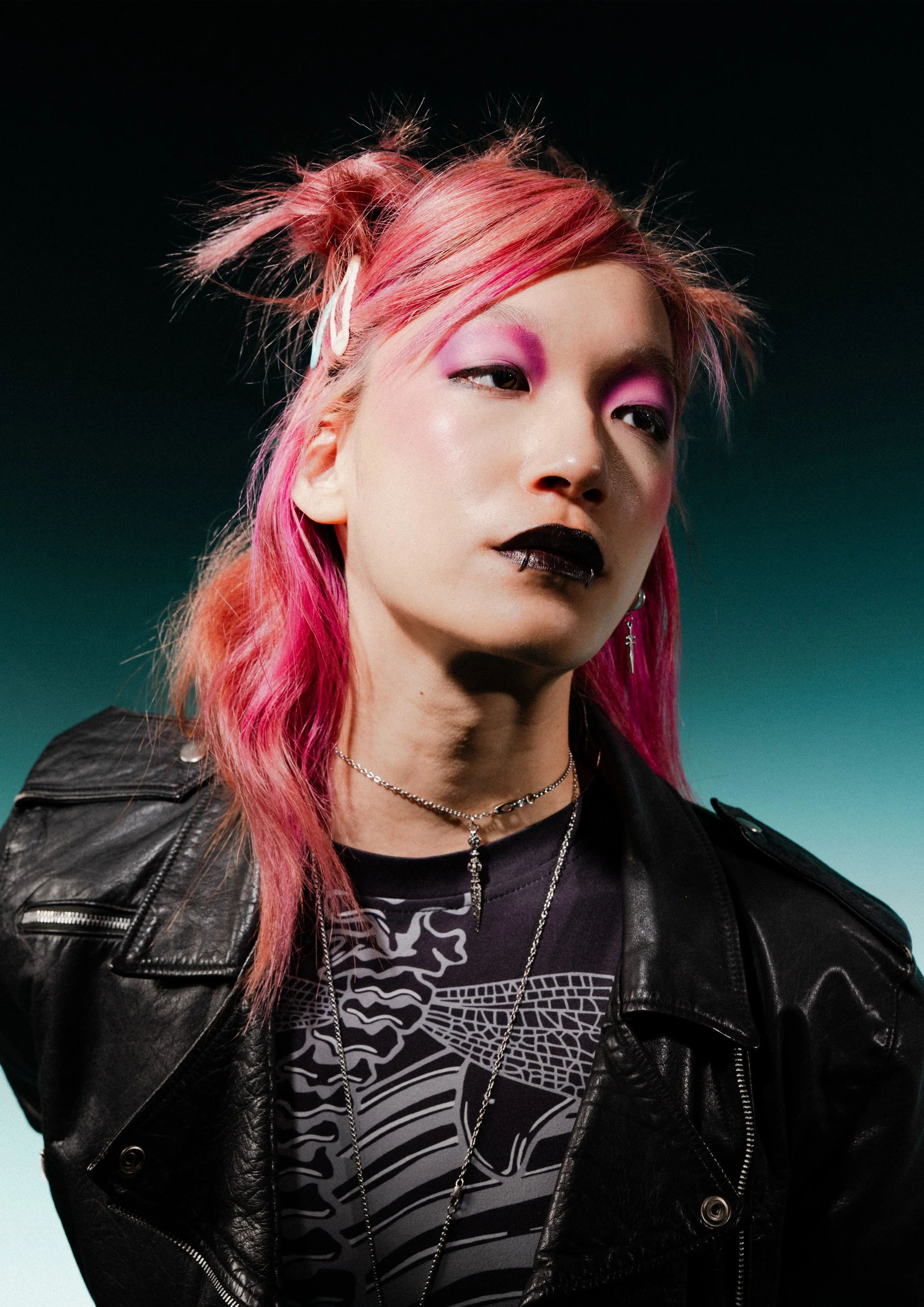 A young woman with pink hair styled in messy buns and teal hair clips, wearing black lipstick, purple eyeshadow, a black leather jacket, a graphic t-shirt, and layered necklaces.