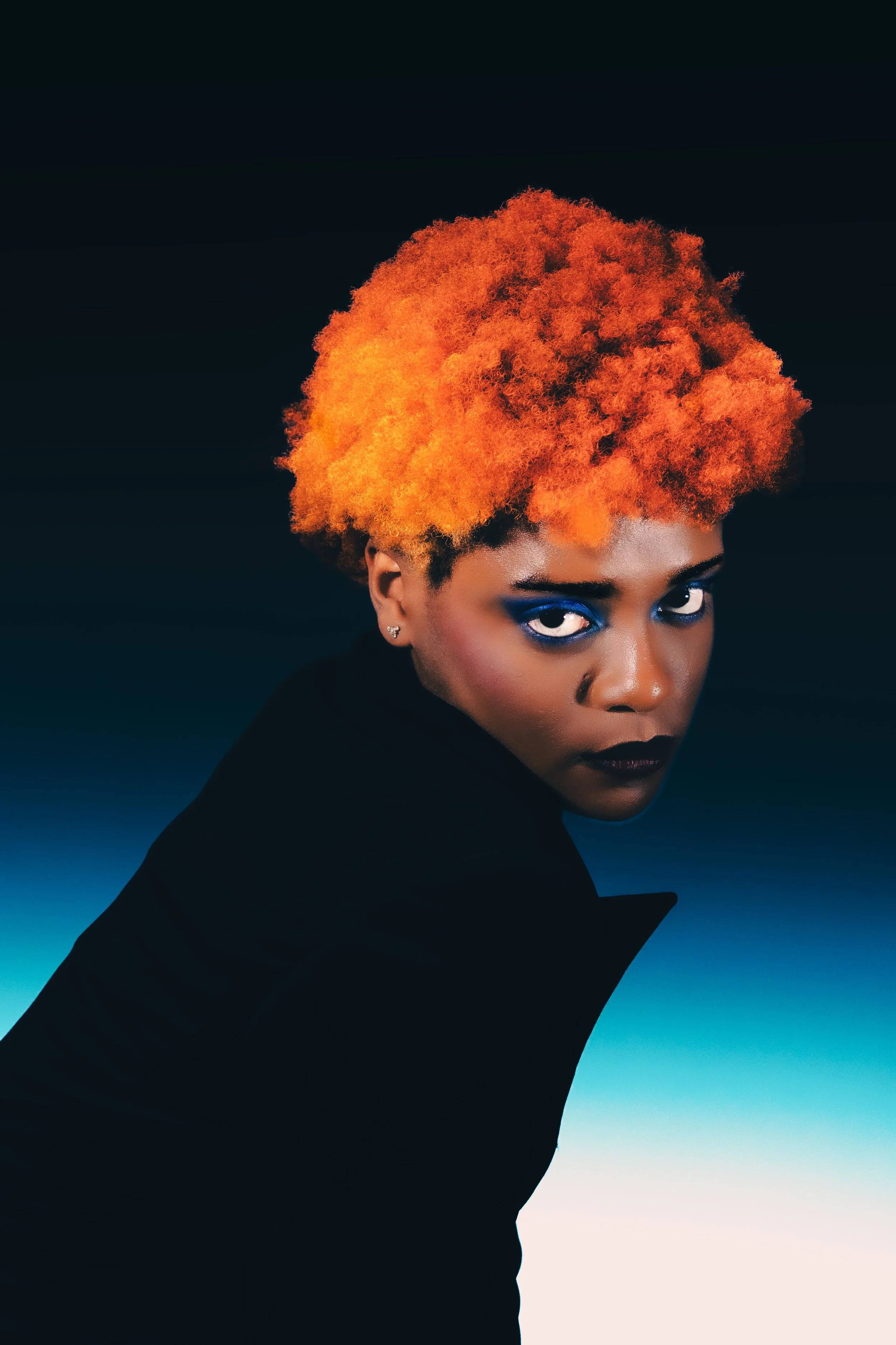 A woman with vibrant orange, curly hair and blue eye makeup looking at the camera against a dark background with a gradient sky.