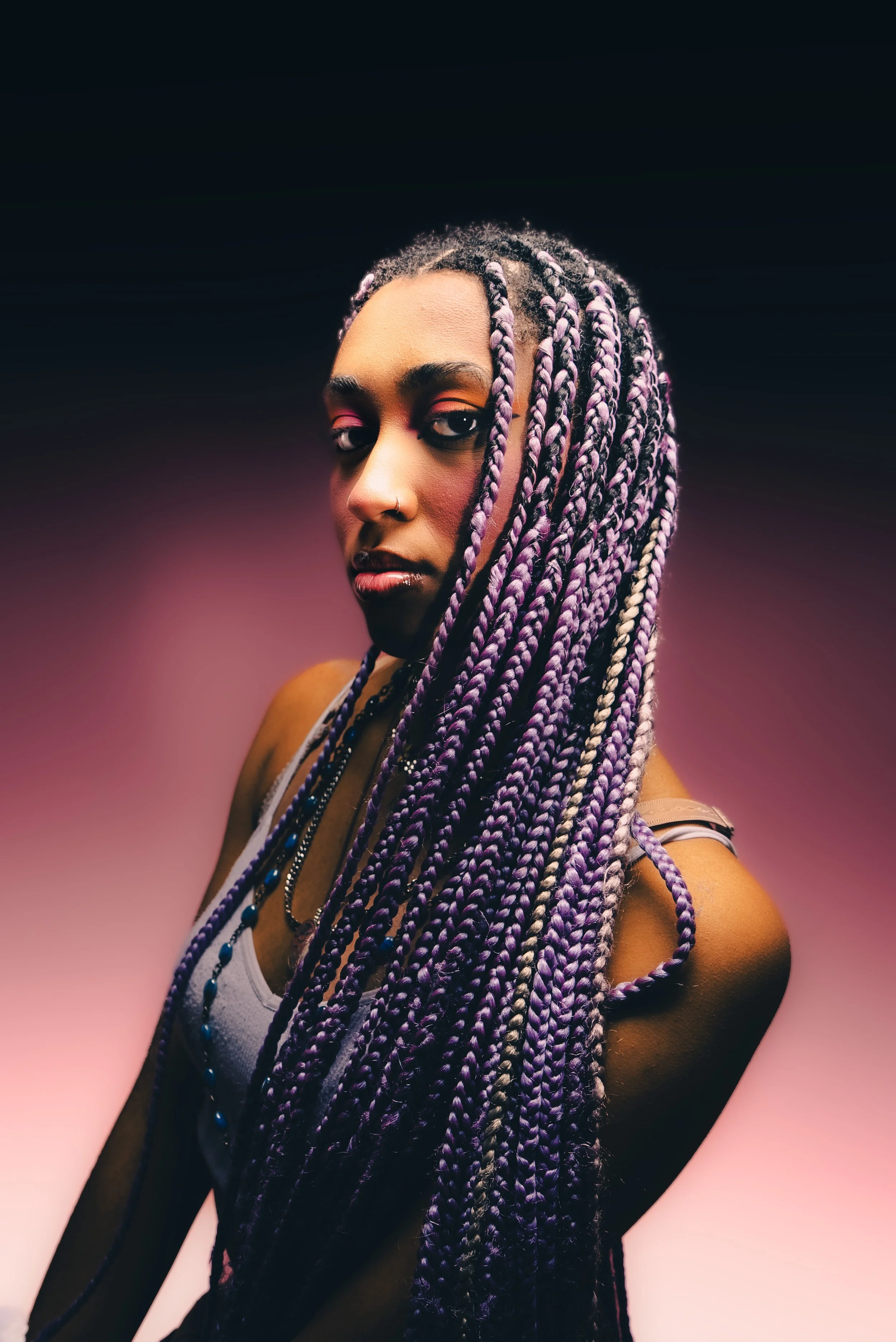 Portrait of a young woman with long purple braids, wearing a nose ring, earrings, and a tank top, against a gradient pink and black background.