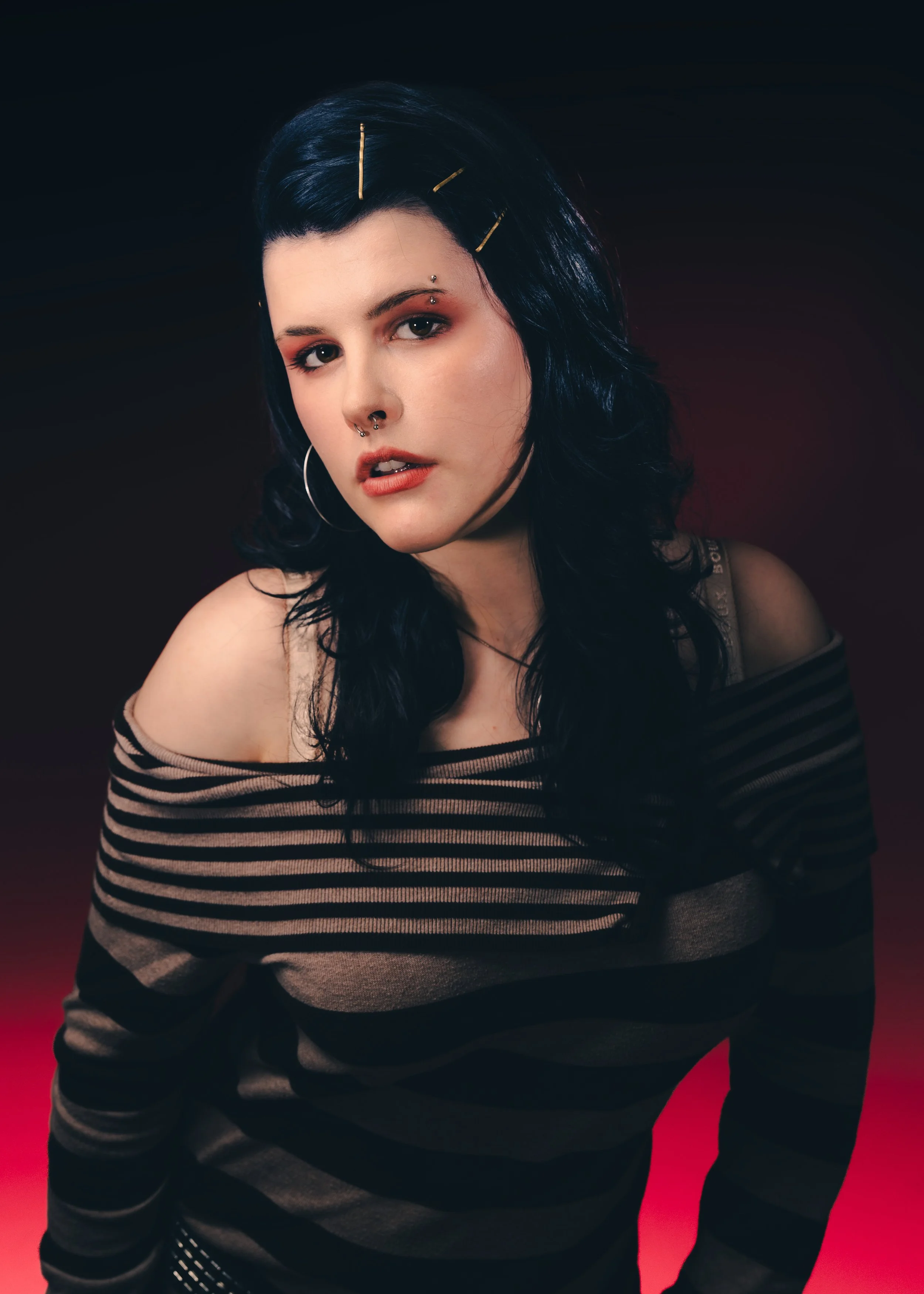 A young woman with black hair and multiple facial piercings, wearing a black and beige striped off-shoulder top, against a dark background with a red floor.