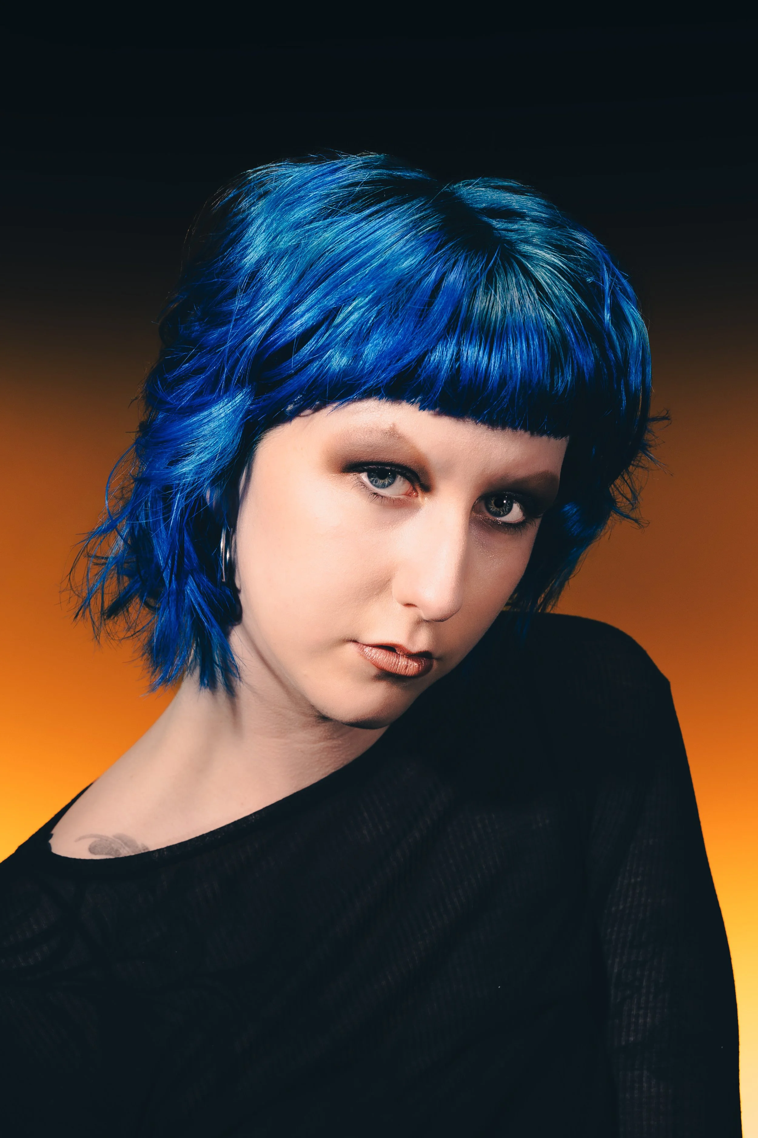 A woman with short, vibrant blue hair and light eye makeup, wearing a black top, against a gradient background transitioning from orange to black.