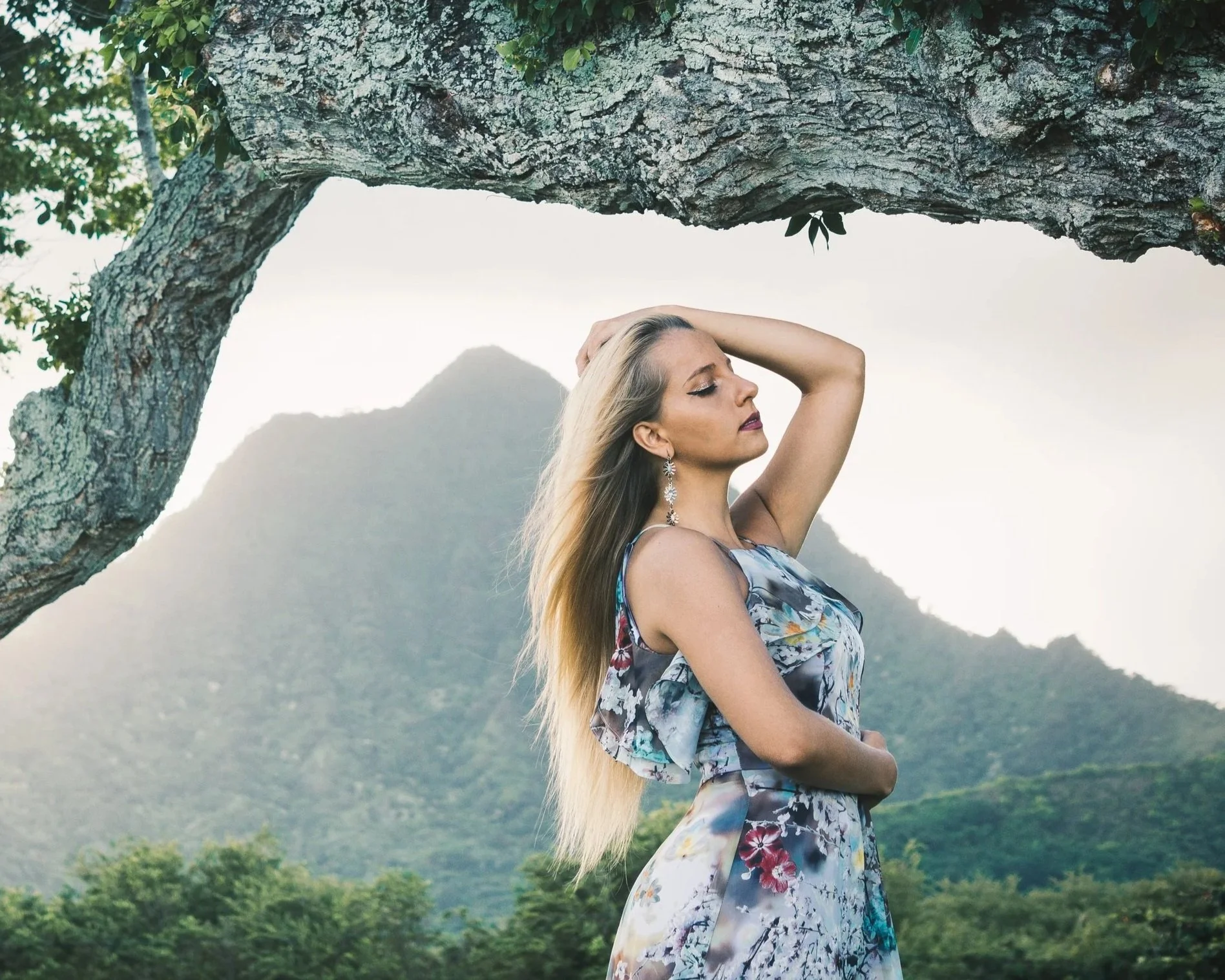 A woman with long blonde hair wearing a floral dress, jewelry, and makeup, is standing outdoors with a mountain and greenery in the background, holding her head with one hand and closing her eyes.