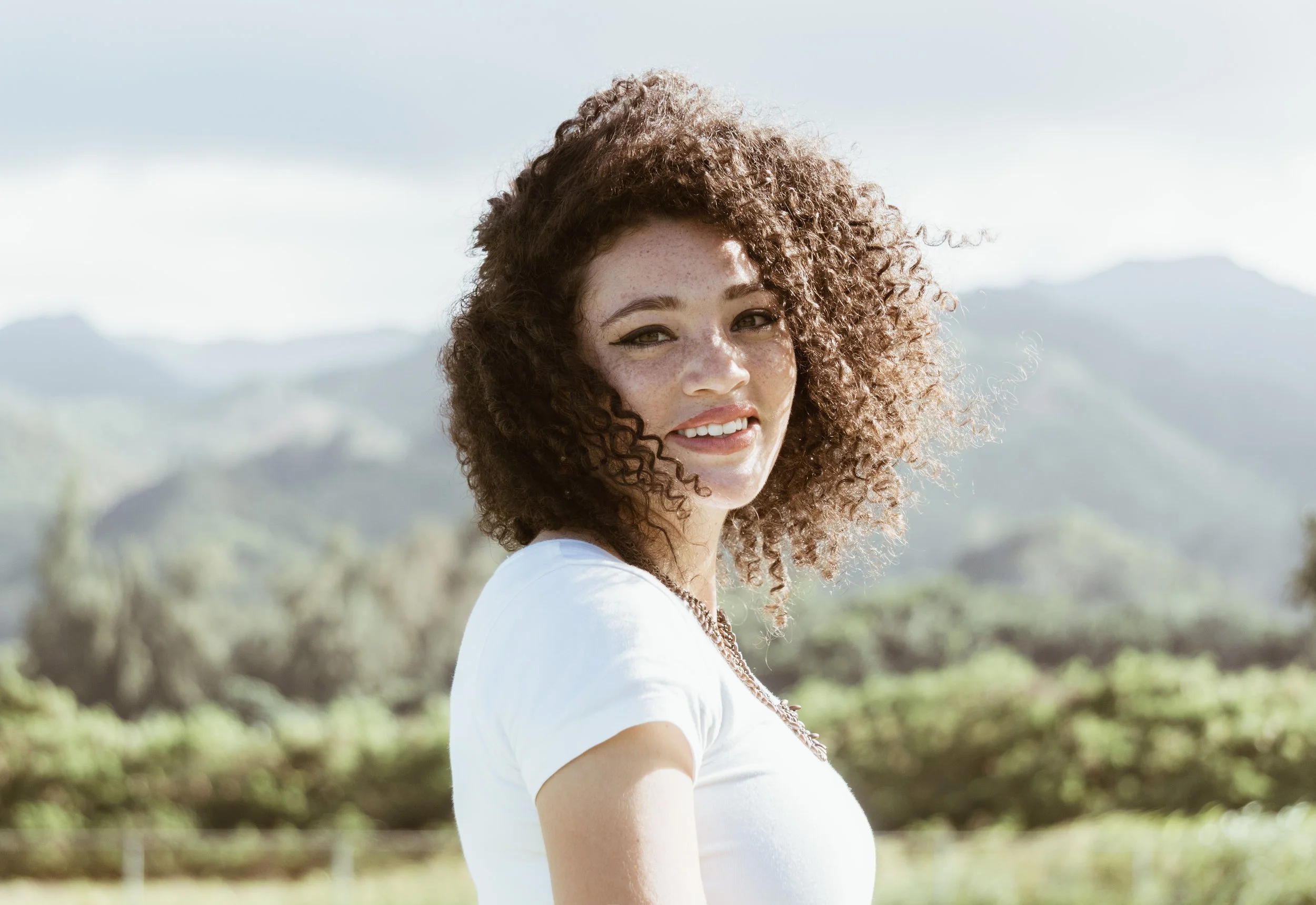 A young woman with curly red hair and freckles, smiling outdoors in a natural setting with mountains and trees in the background.