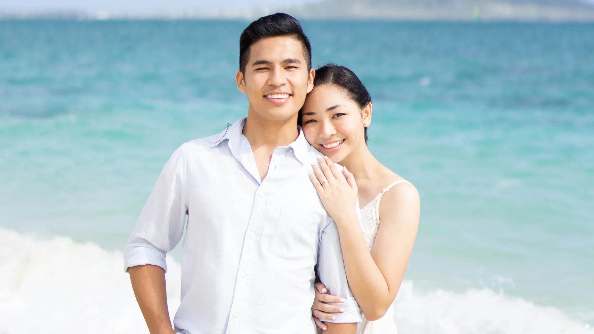A smiling young man and woman standing close together on a beach, with the ocean and waves in the background.
