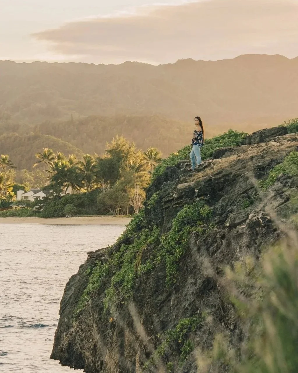 A woman standing on a rocky cliff overlooking the ocean with a hillside and trees in the background during sunset.