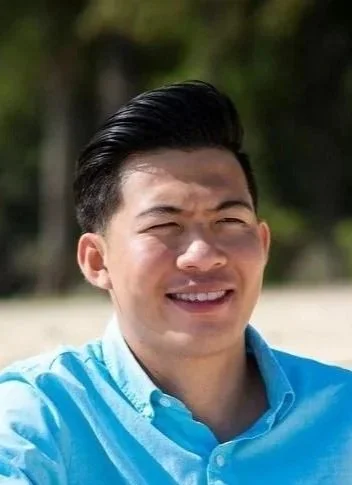 A young man smiling outdoors with a background of trees and a beach, wearing a light blue shirt.