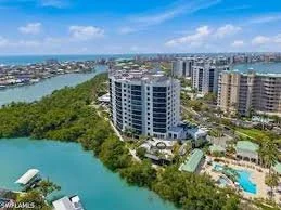 Aerial view of a south end of Fort Myers Beach with tall residential buildings, the Estero Back Bay, and lush green trees, with a swimming pool visible among the buildings and a bright blue sky overhead.