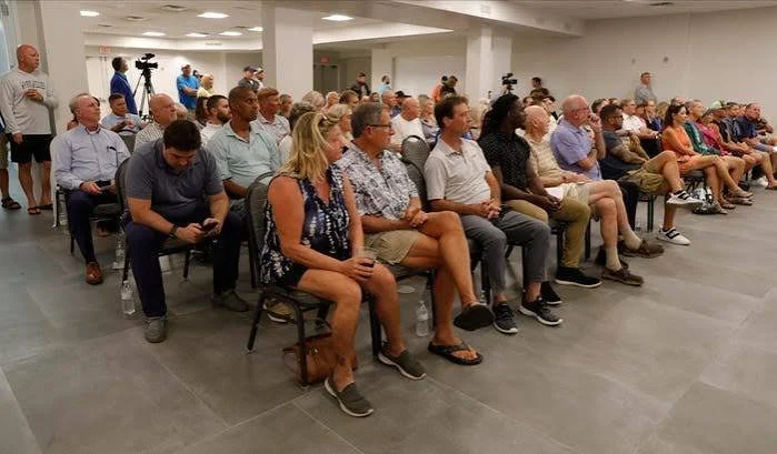 Audience seated in rows at a Fort Myers Beach community forum with some media personnel and cameras in the background.