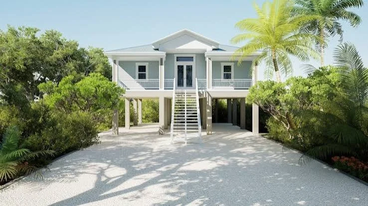 Beach house on stilts surrounded by tropical trees with sandy path leading to the door in a sunny setting.