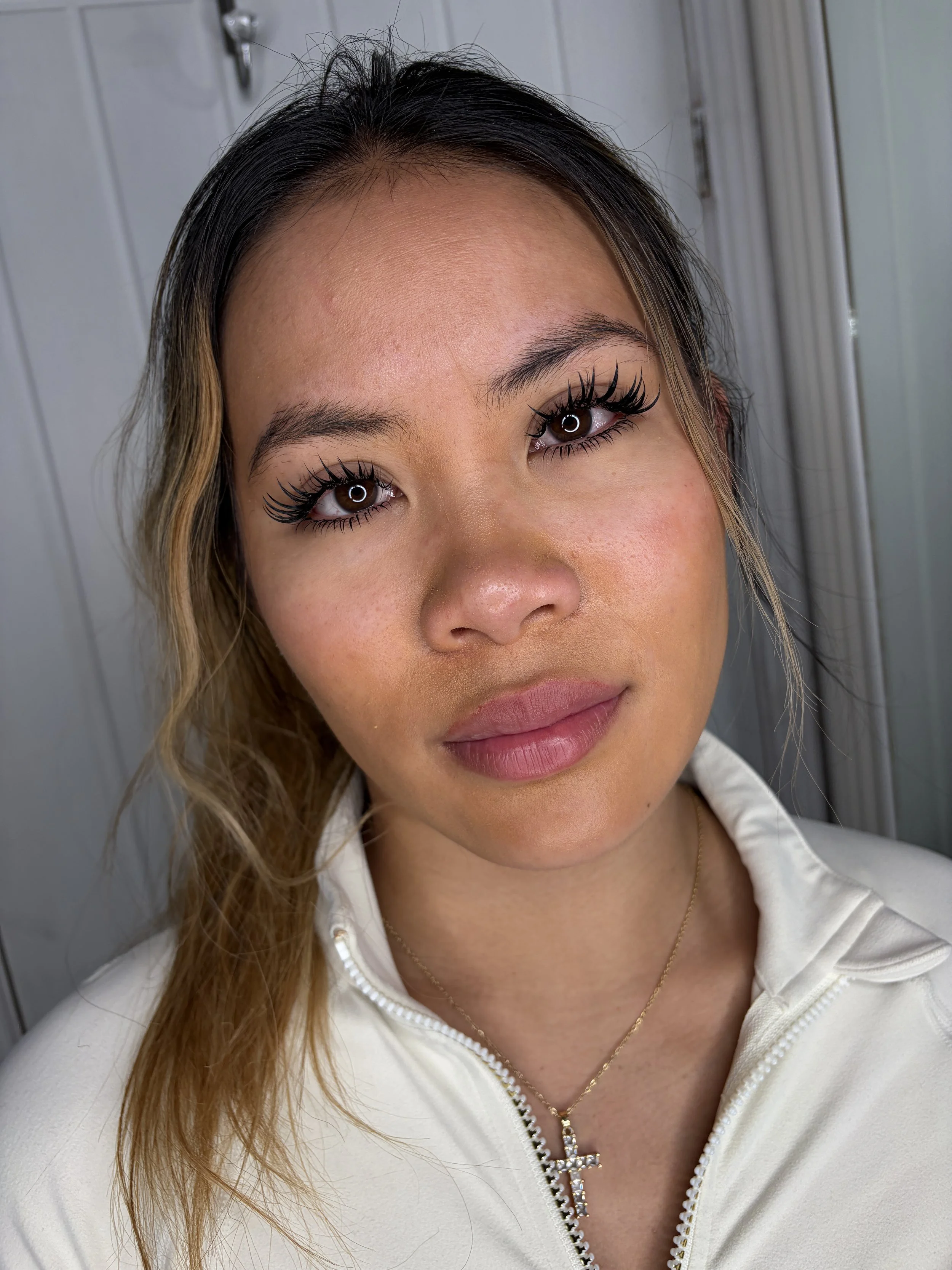 Close-up of a woman with long wavy hair, wearing makeup including long false eyelashes, a white zip-up hoodie, and a gold necklace with a cross pendant.
