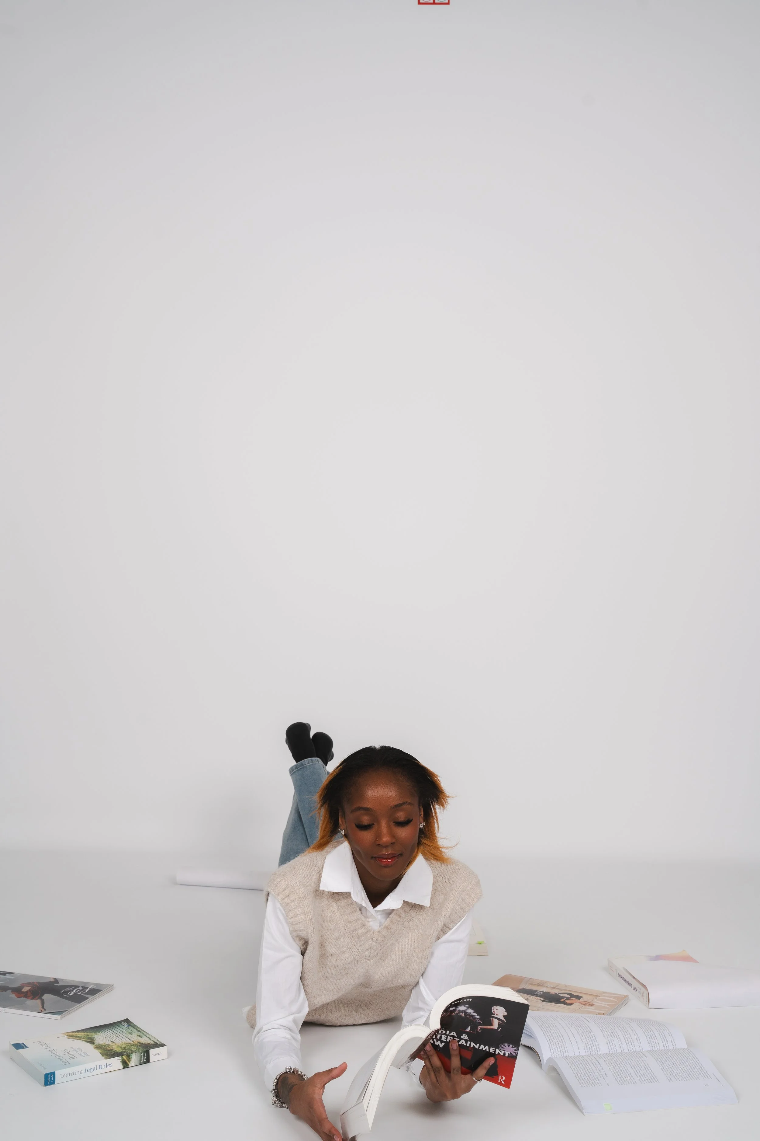 A woman with short hair and earrings is sitting at a table, reading a magazine with a film theme, while other magazines and books are spread on the table. Behind her, a person with legs up on the table is hanging upside down, wearing a denim jacket, black gloves, and no shoes against a plain white background.