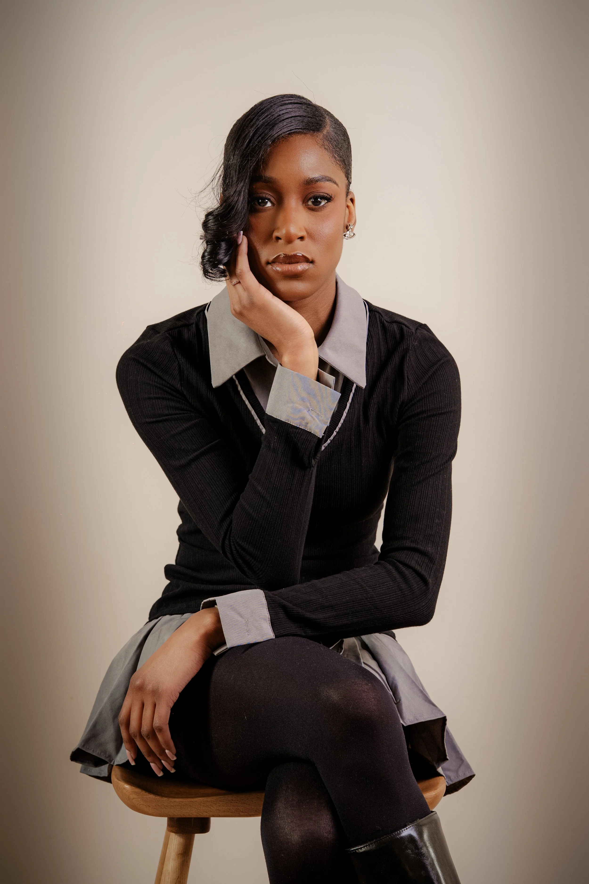 A young woman with short, styled black hair, sitting on a wooden stool against a plain beige background, wearing a black sweater with gray cuffs, a gray skirt, black tights, and shiny black boots, posing with her chin resting on her hand.