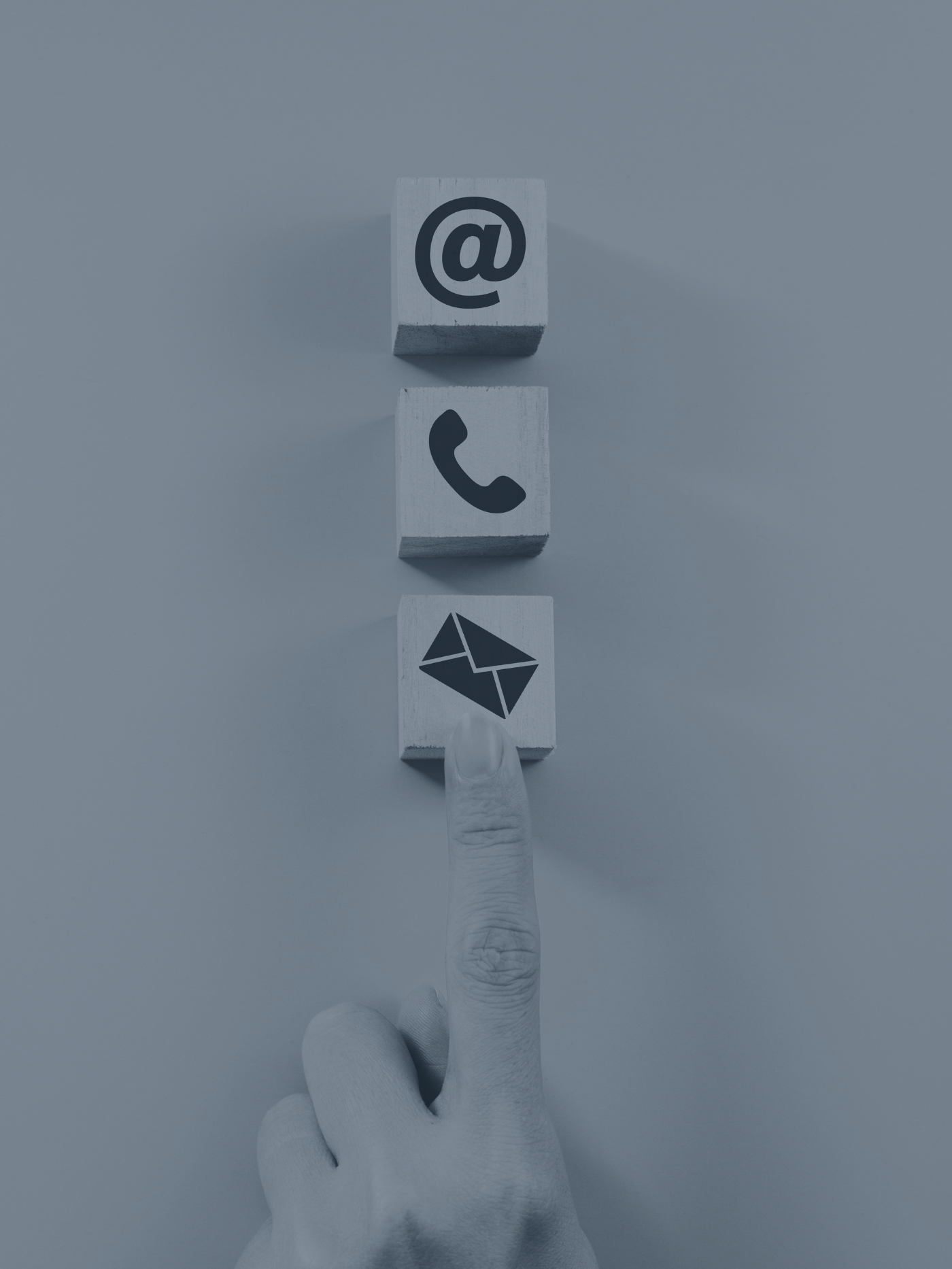 A person's hand pointing at three small wooden blocks on a white surface. The blocks display social media icons for email, phone, and at symbol.