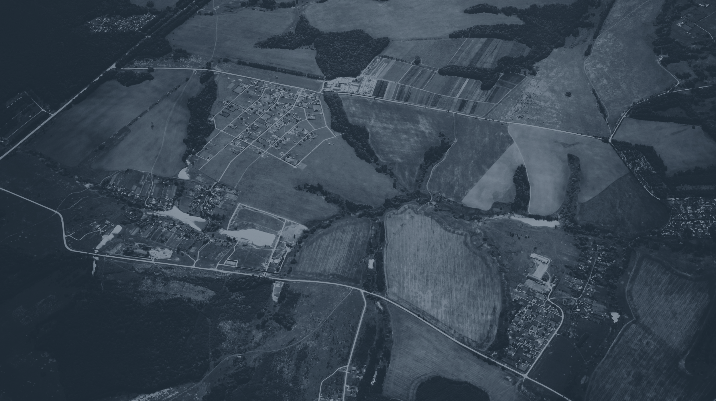 An aerial view of a rural area with fields, roads, and small clusters of buildings, surrounded by patches of forest.
