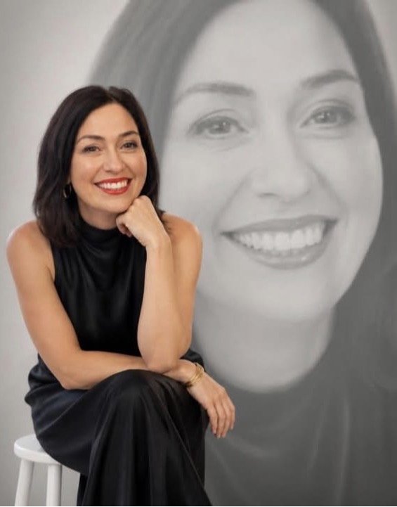 A woman with dark hair, wearing a black sleeveless dress, smiling and sitting on a white stool in front of a large black-and-white portrait of herself.