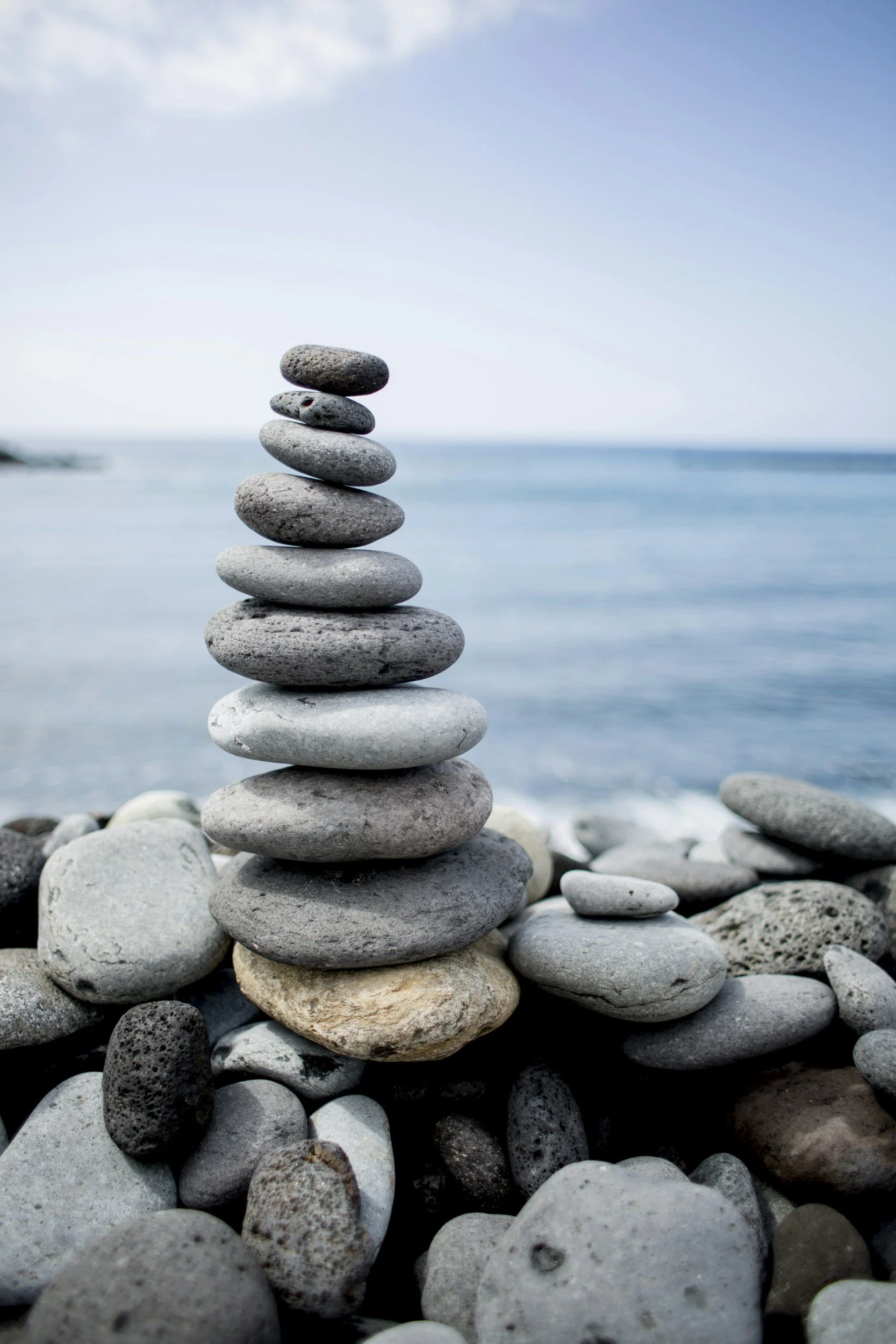 A stack of seven smooth, rounded stones balanced on top of each other on a pebble-covered beach with the ocean and a partly cloudy sky in the background.