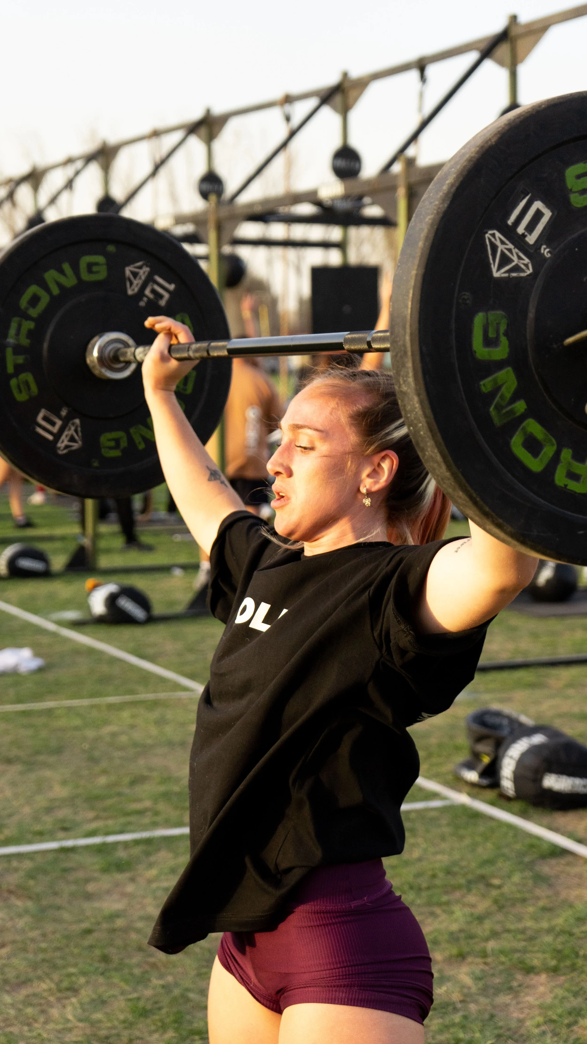 A woman lifting a barbell overhead during an outdoor workout at sunset, with gym equipment and people in the background.
