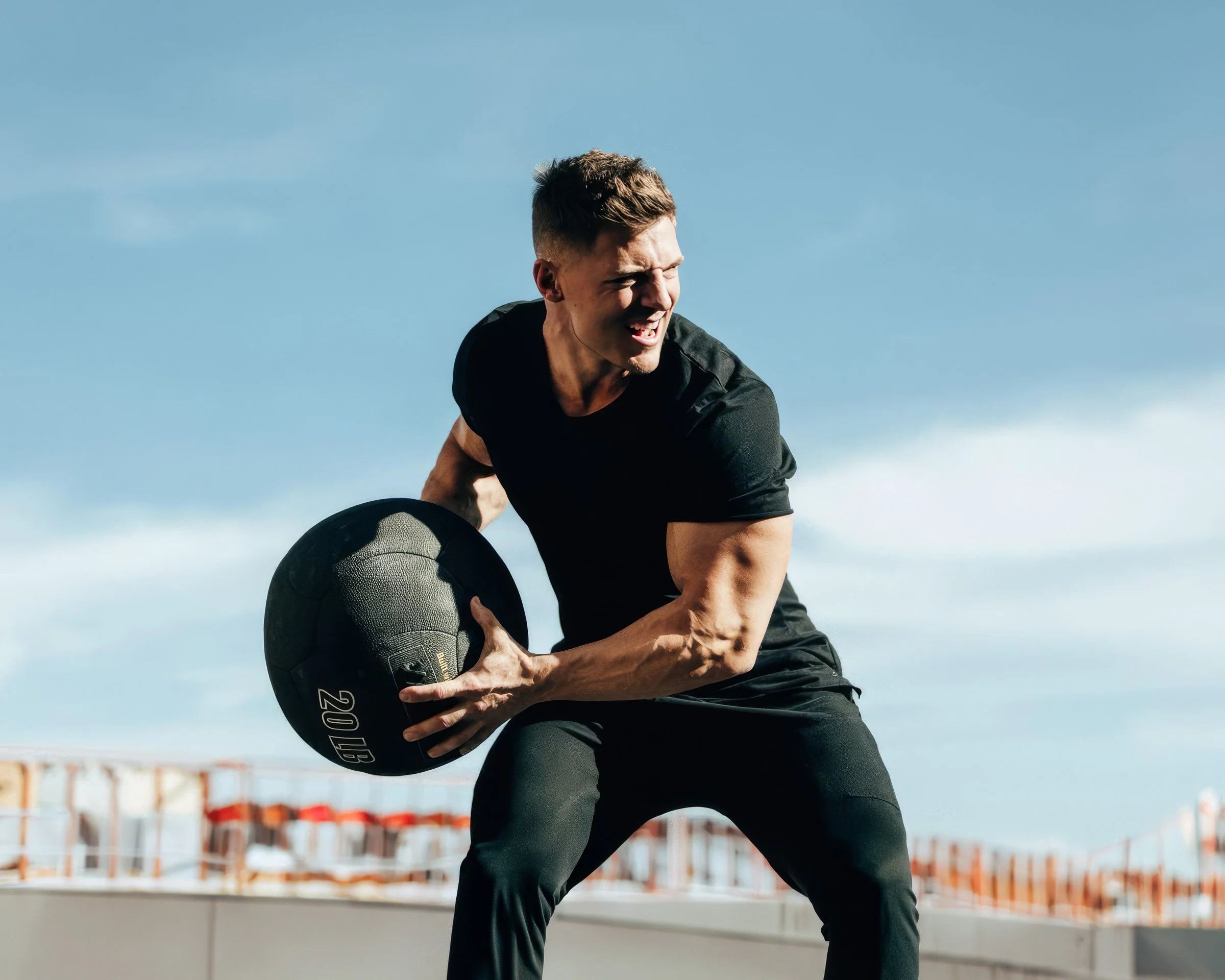 A man with short dark hair and a serious expression, wearing a black t-shirt, is outdoors against a clear blue sky. He is holding a black medicine ball labeled '20 LB' with both hands, preparing for a workout or exercise. The background shows a construction site with scaffolding and orange safety barriers.