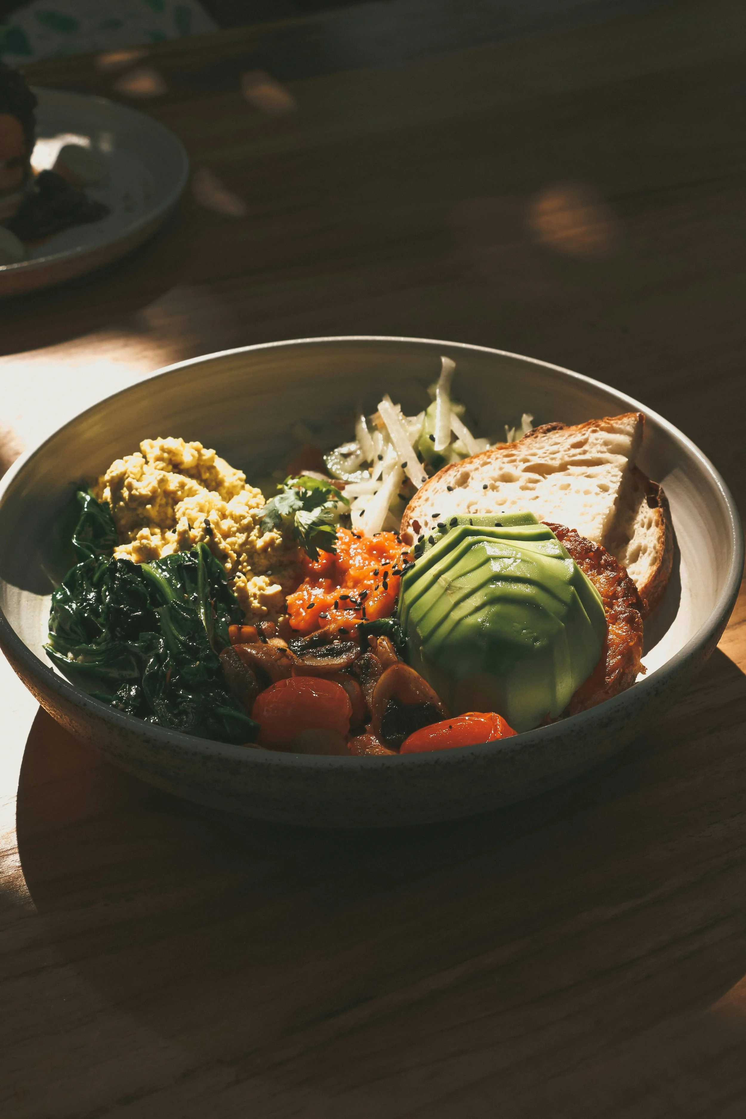 A bowl of breakfast food including sliced avocado, scrambled eggs, cooked greens, cherry tomatoes, and bread.