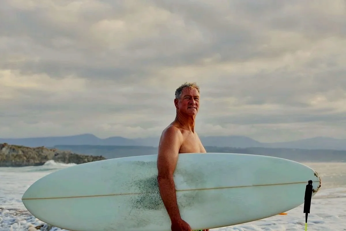 Older man with gray hair holding surfboard on the beach with cloudy skies and mountains in the background.