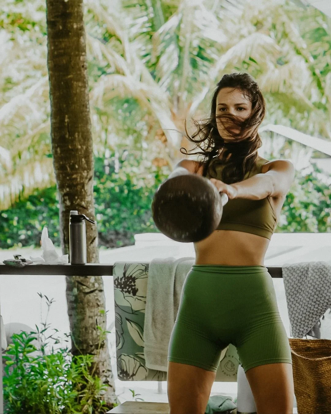 A woman with long hair wearing a brown sports bra and green high-waisted shorts is exercising outdoors with a kettlebell. She is standing with her arms extended forward, gripping the kettlebell, with a background of lush green tropical plants and trees.