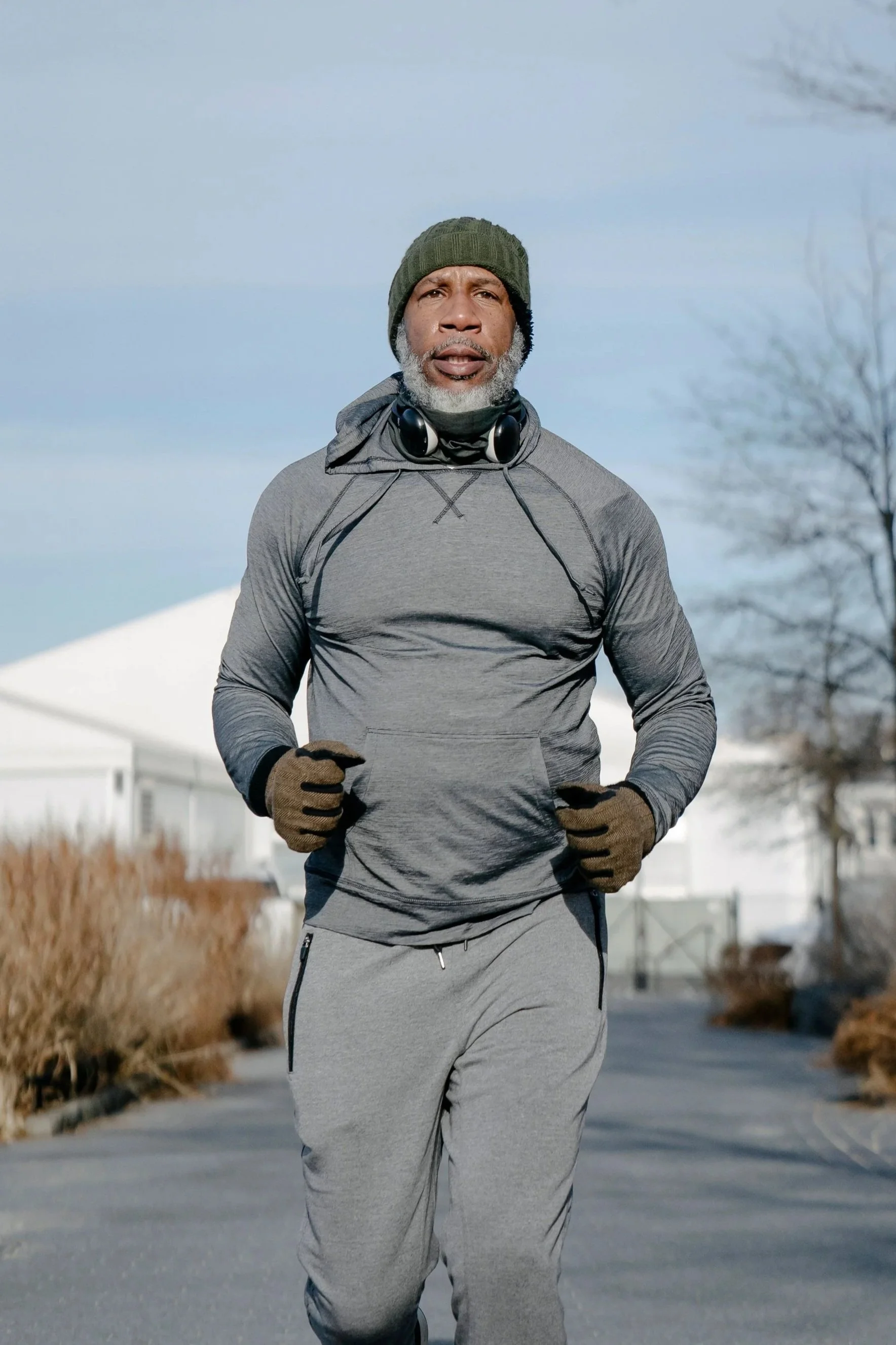 An elderly man running outdoors in winter, wearing a green beanie, gray athletic hoodie, gloves, and gray sweatpants, with a serious expression and snow-covered ground.