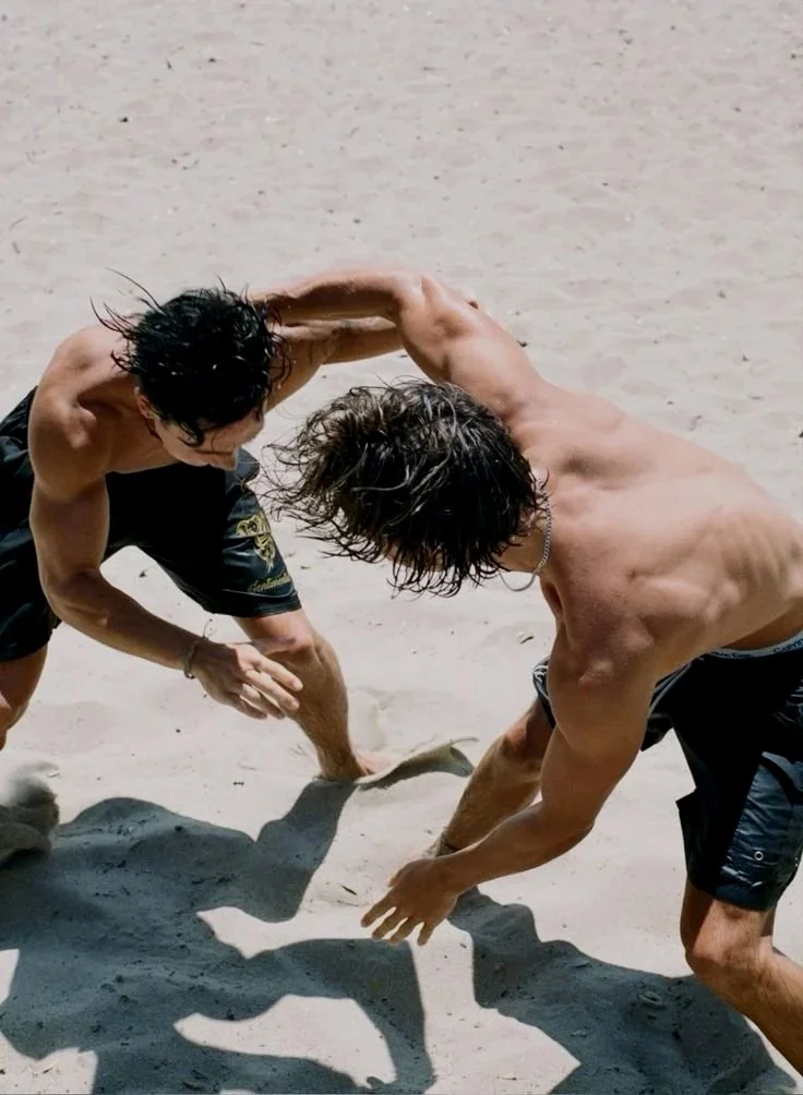 Two shirtless men wearing black shorts playing a game of beach football on sand.