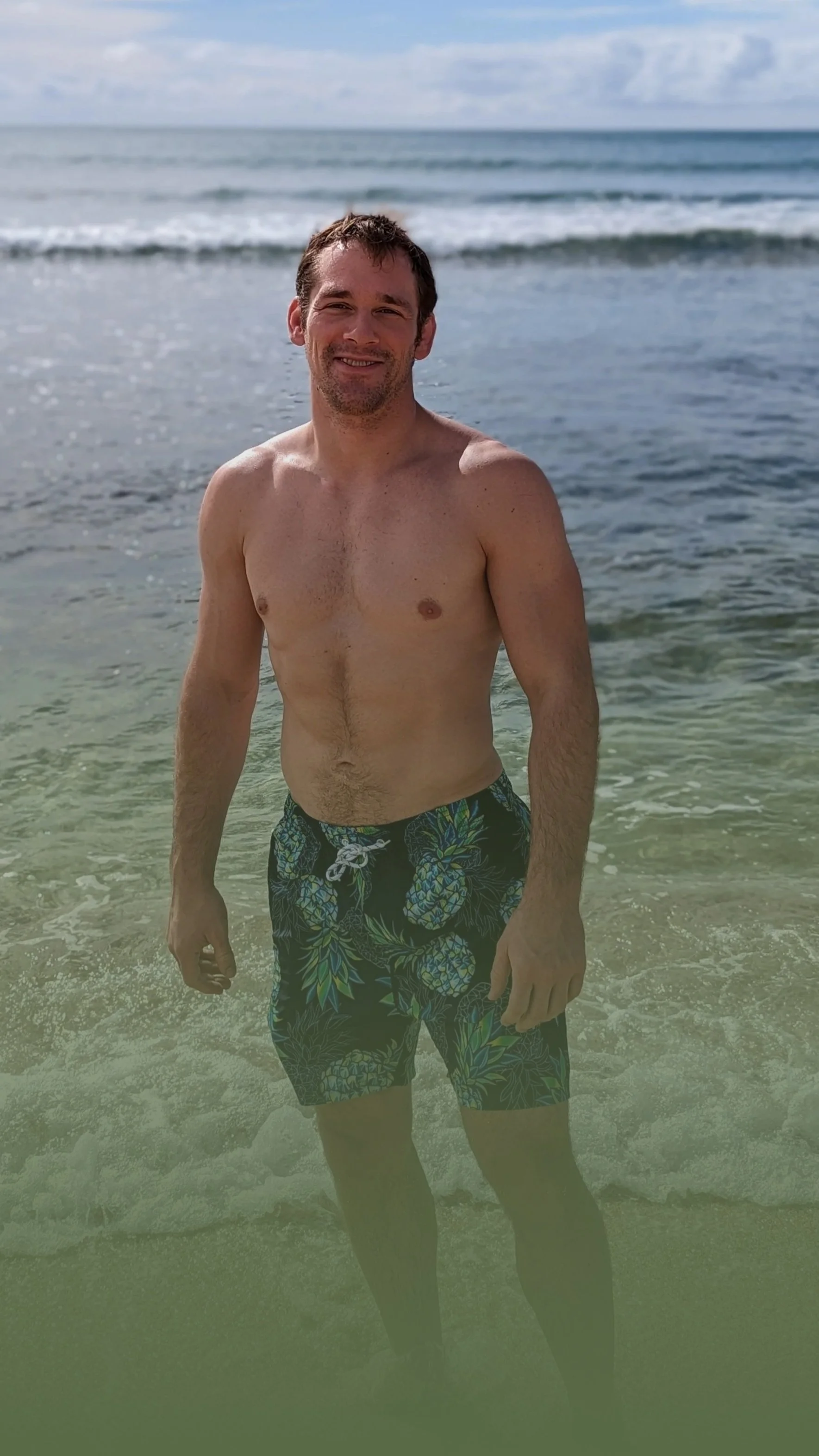 A man standing in the ocean waves at the beach, wearing black shorts with a pineapple design, smiling with wet hair.