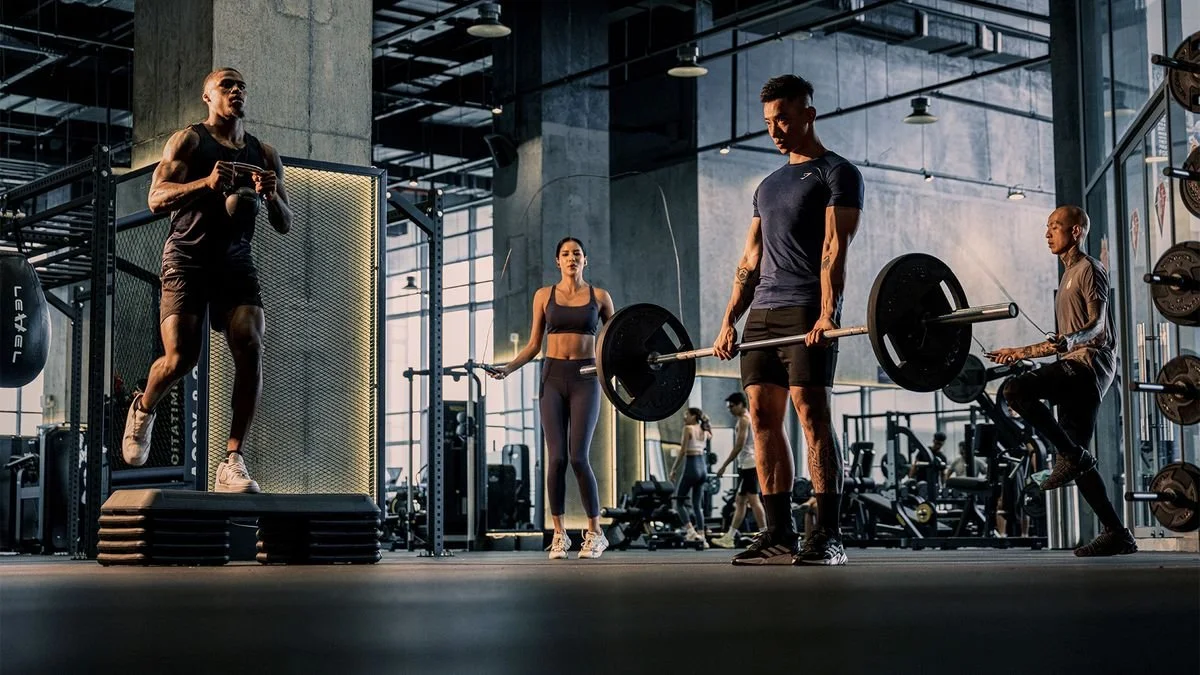 People working out in a modern gym, including a woman performing box jumps, a man lifting a barbell, two women jumping rope, and gym equipment in the background.