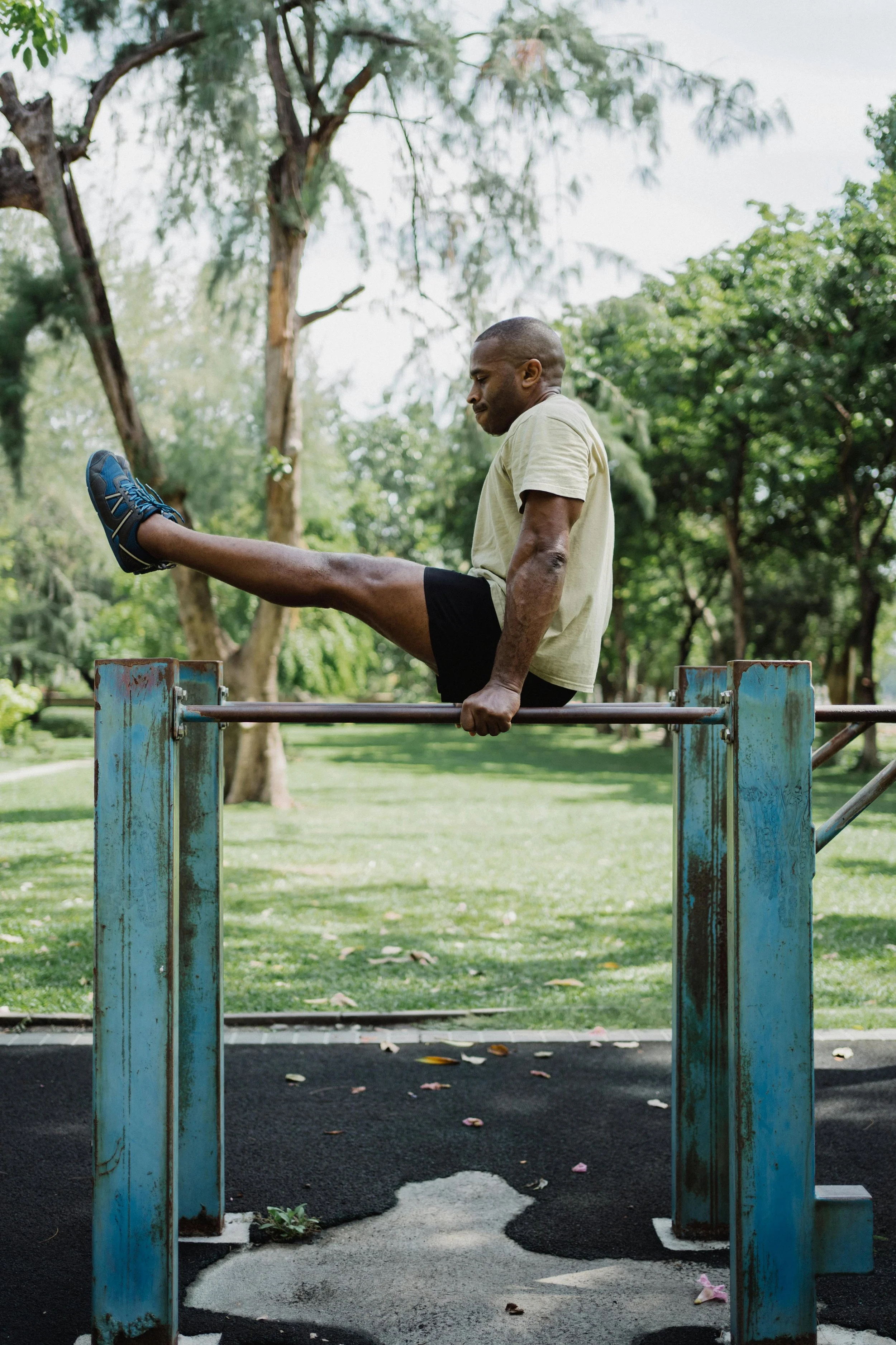 A man performing a parkour move by balancing on a horizontal bar in a park with green trees and grass in the background.