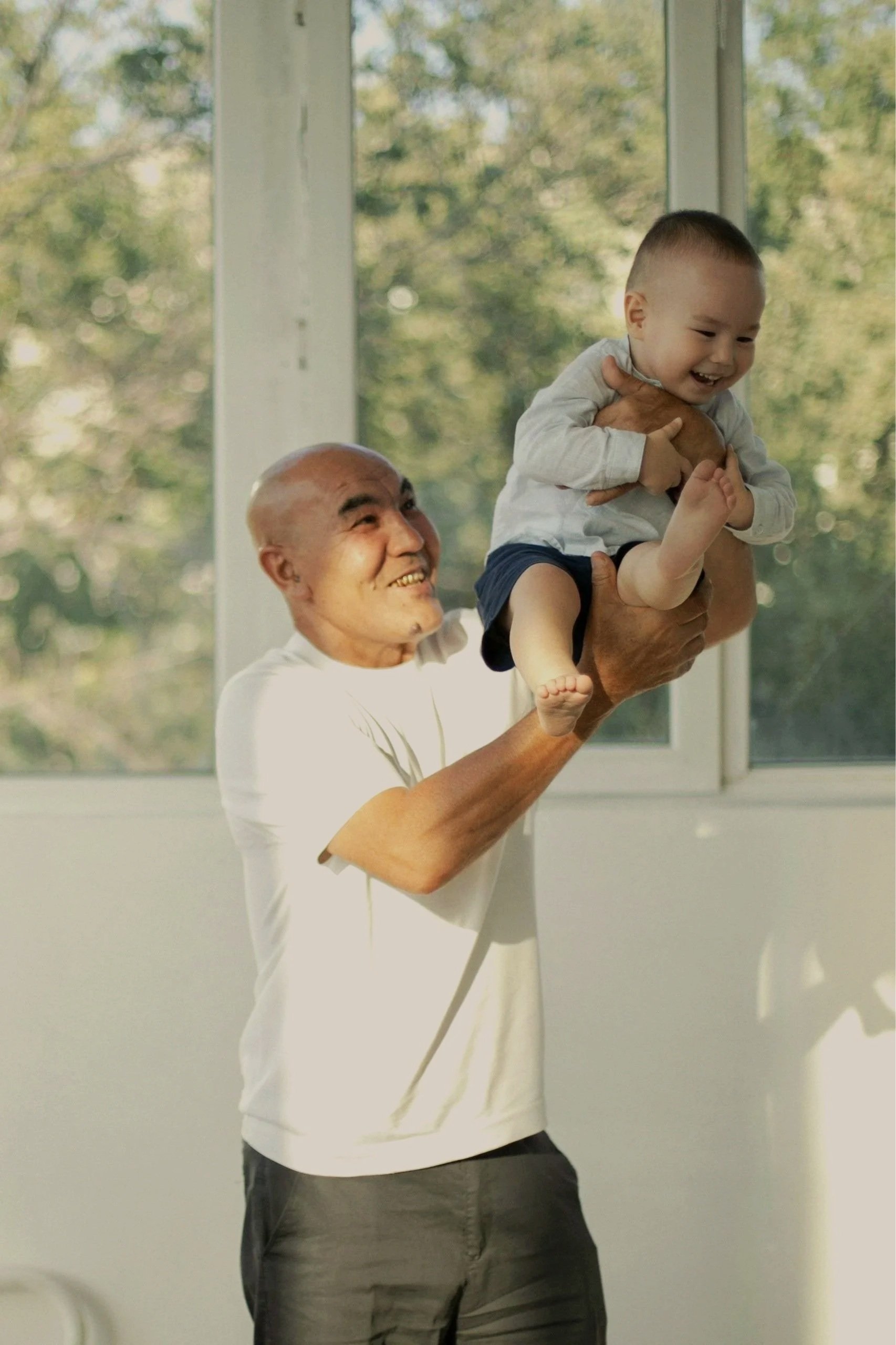 An elderly man with a shaved head holding a young boy with a short haircut and a big smile in front of a window with trees outside.