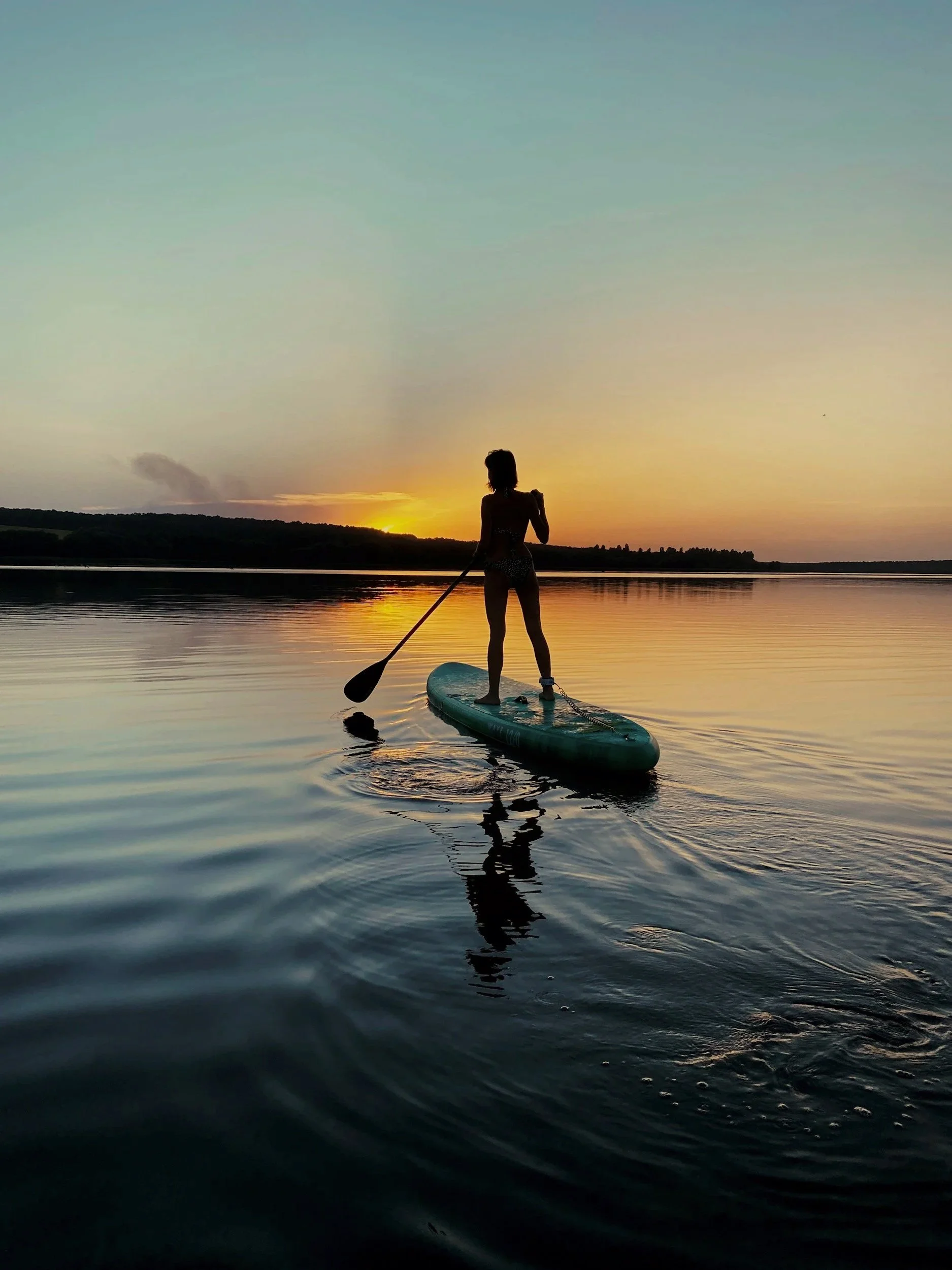 Person paddleboarding on a calm body of water during a sunset, with a silhouette of hills in the background.