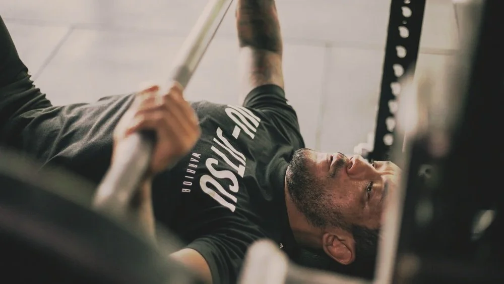 Man lifting weights in gym, lying on a bench press, wearing a black t-shirt with white text.
