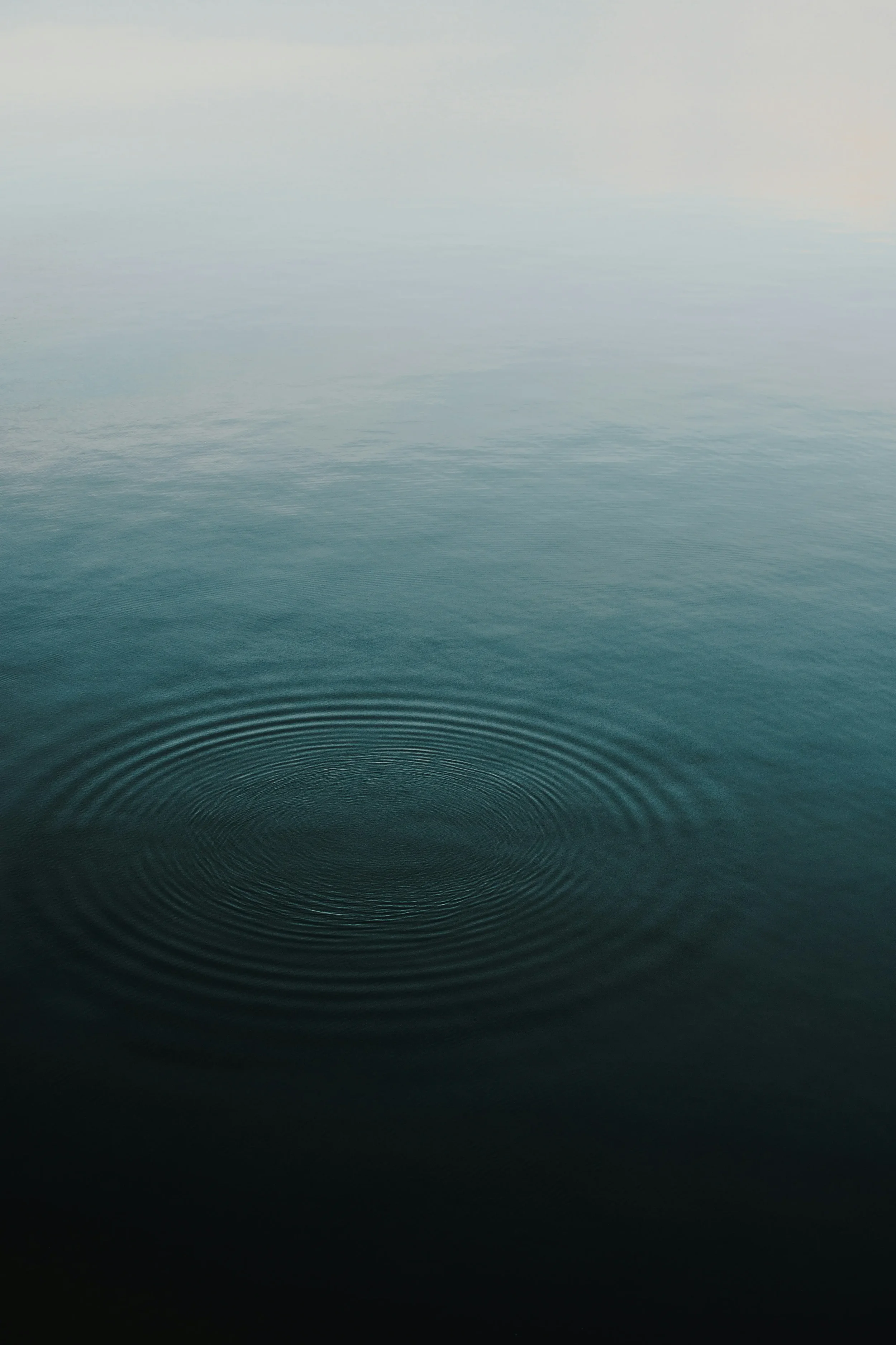 Calm body of water with concentric ripples in the foreground, gradient shades from dark to light blue with foggy sky in the background.