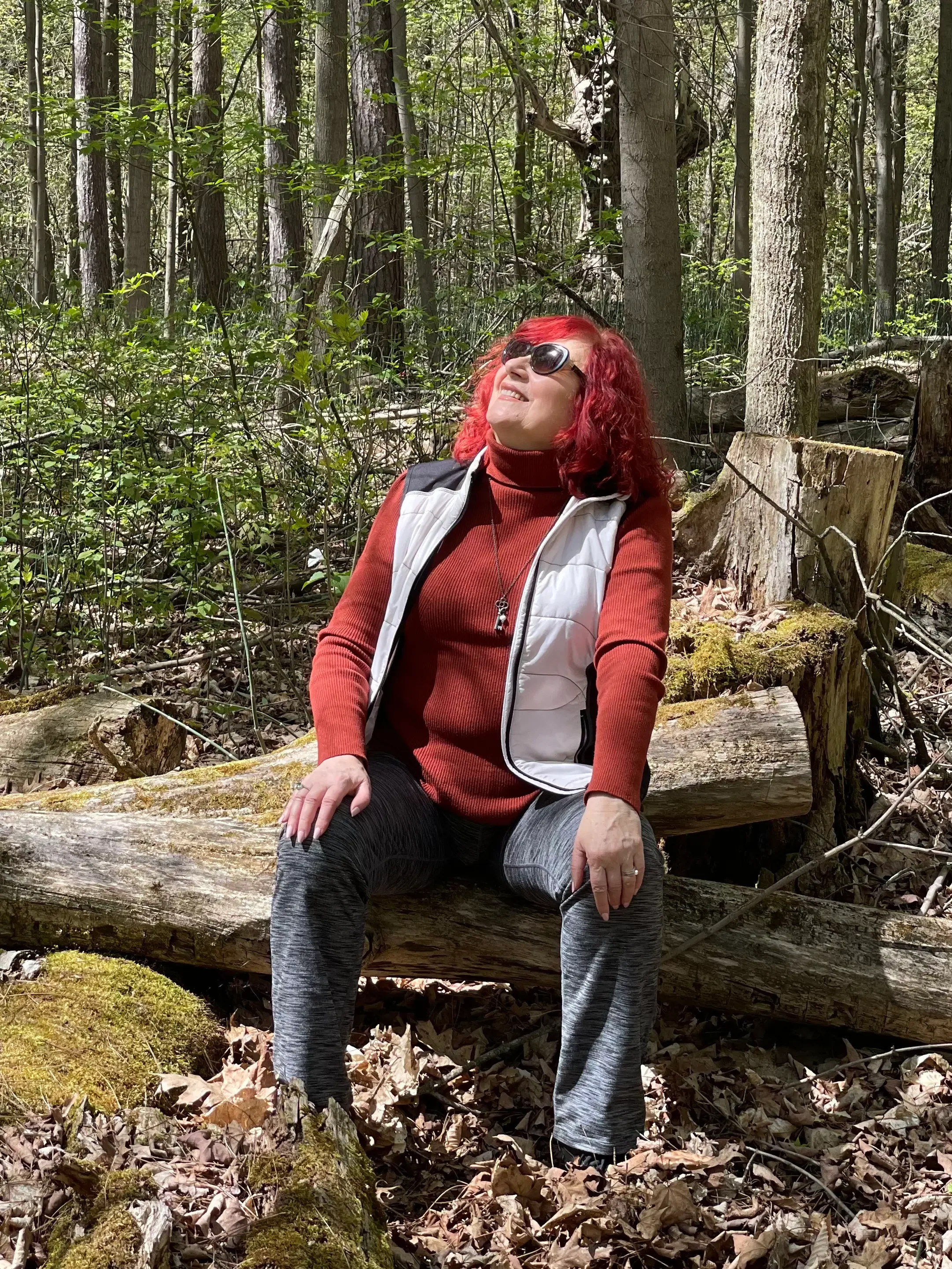 Photo of woman in red sweater and white vest with red hair and sunglasses sitting on a fallen tree in the woods.