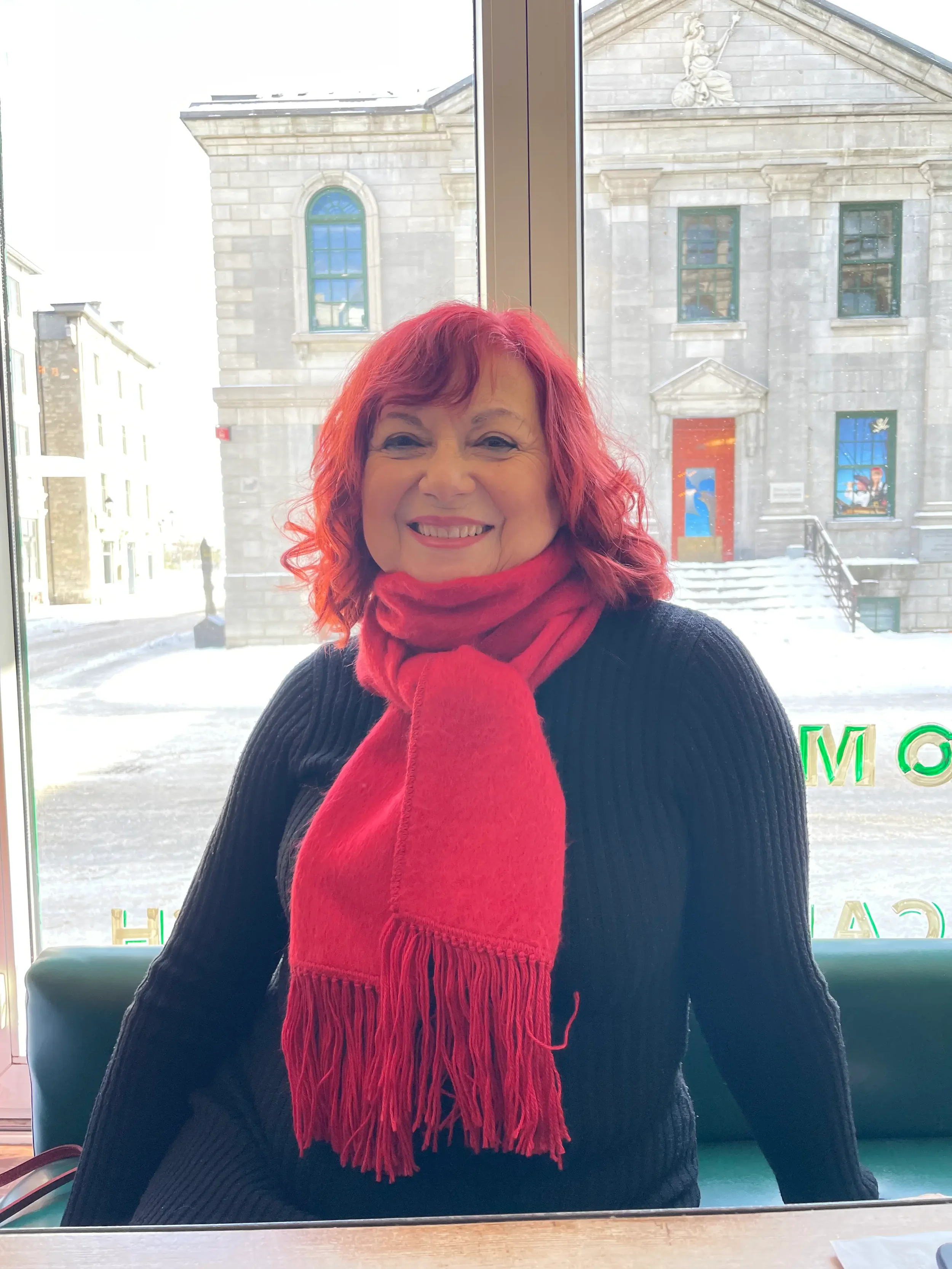 A woman with red hair and a red scarf smiling inside near a window with a snowy street view and a stone building outside.