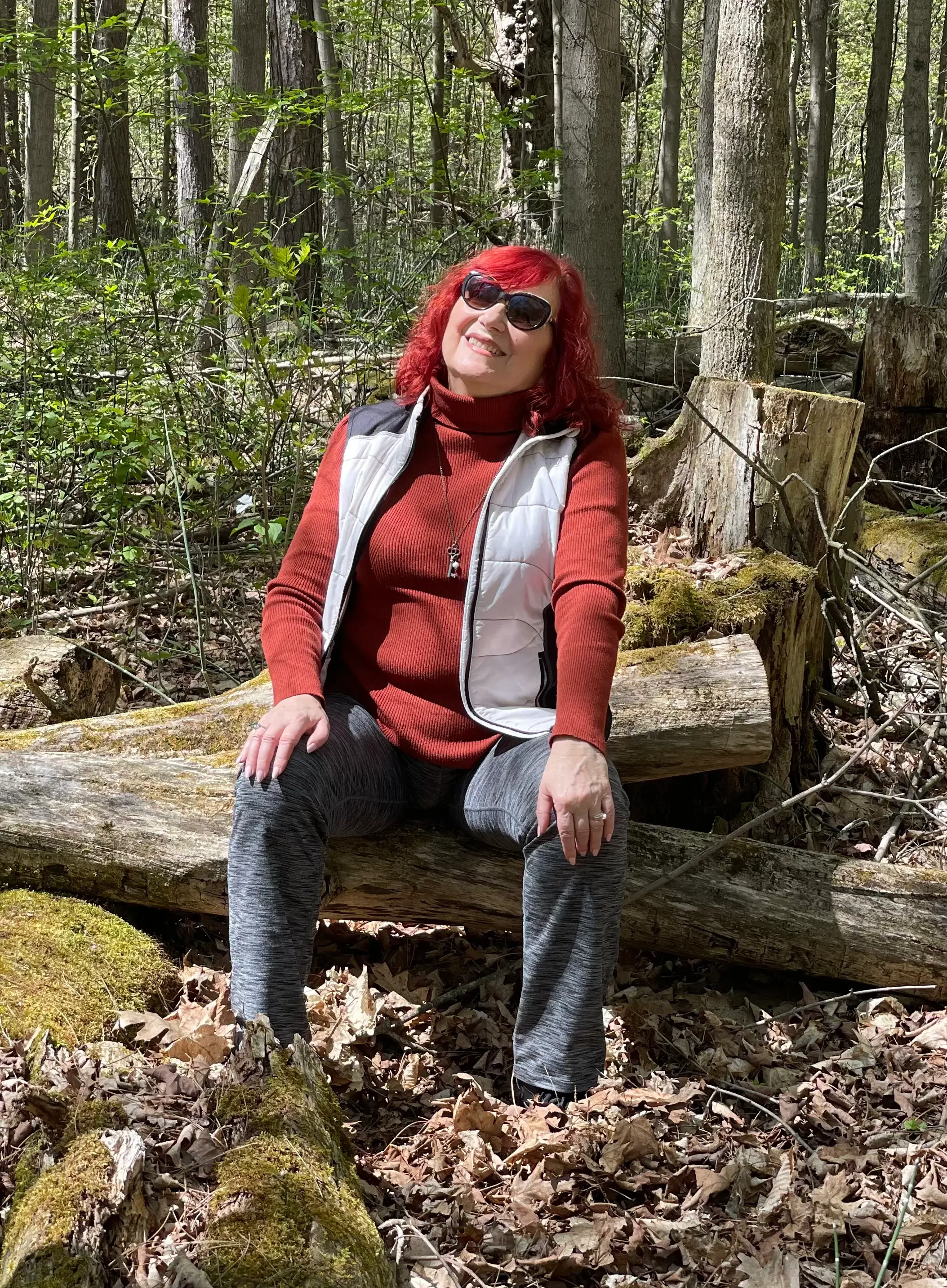 Photo of woman in red sweater and white vest with red hair and sunglasses sitting on a fallen tree in the woods.
