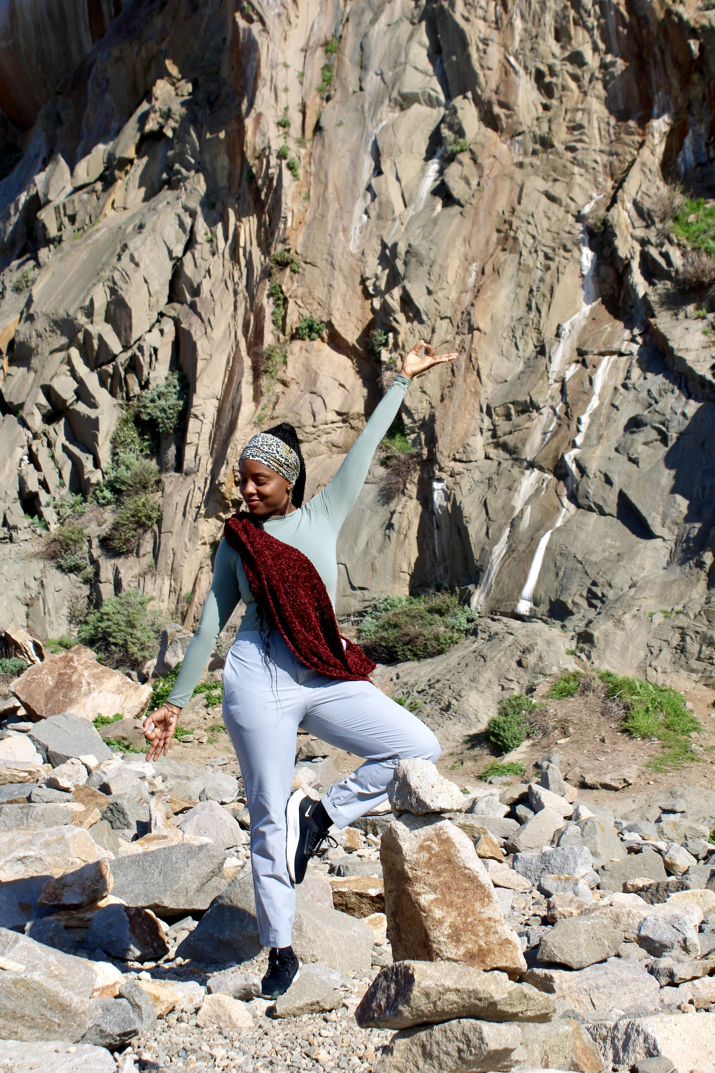 A woman practicing yoga outdoors among rocks and a rocky cliff, wearing a headband, long-sleeve shirt, and white pants, balancing on one leg with arms raised.